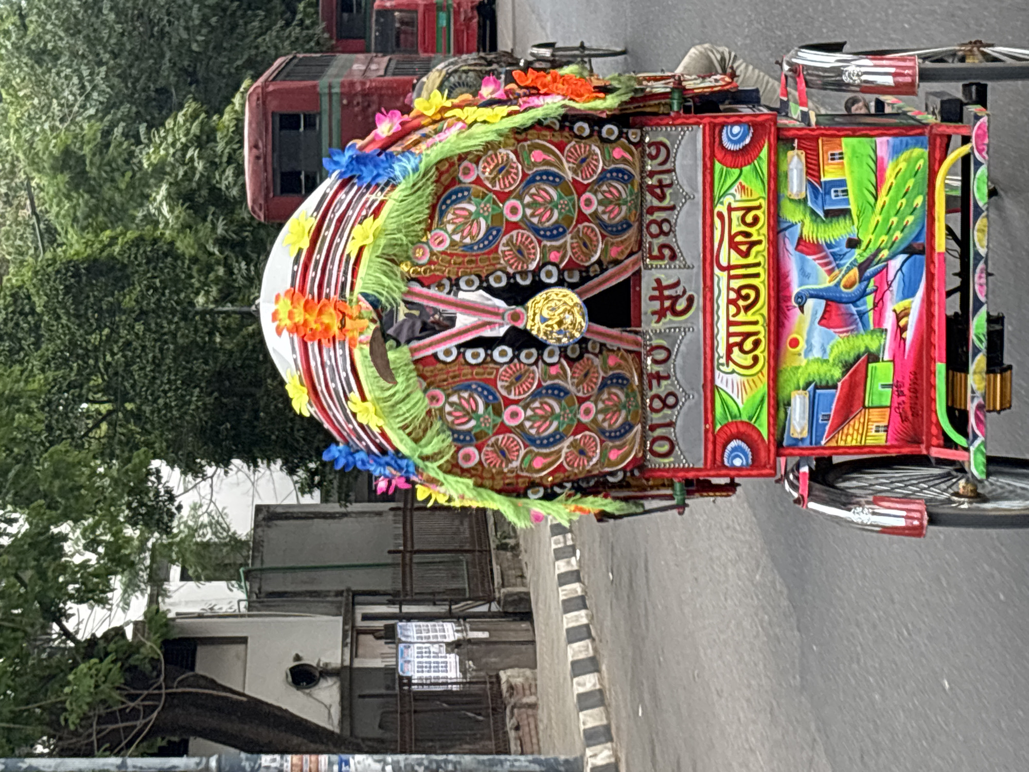 A vibrant, decorated rickshaw stands on a street in Dhaka, Bangladesh. It is adorned with colorful floral motifs, a depiction of a peacock amidst houses, and the Bengali word "Moshakhin" painted vertically, along with the number "01870-581419" below in smaller writing. The rickshaw also features neon green fringe and colorful faux flowers. The street is paved, with a curb painted in black and white stripes. Buildings are visible in the background, along with trees lining the street. A red bus is visible in the background with the bus's lights illuminated.