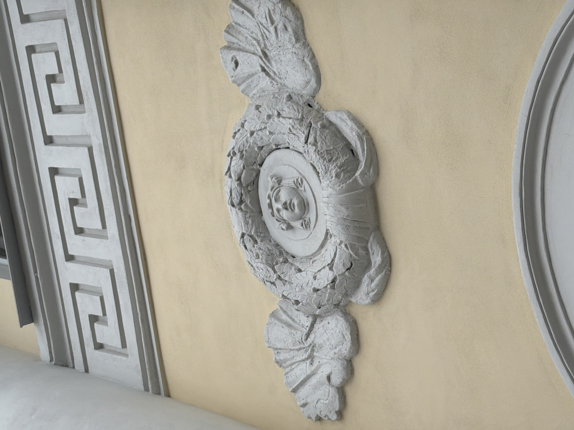 The image depicts an ornate, grey bas-relief architectural element affixed to a light yellow wall. The design includes a central circular medallion featuring a face in profile, surrounded by sculpted leaves or foliage. Above and below the medallion are stylized leaf or floral motifs. To the left, a rectangular border with a Greek key pattern is visible. To the right, a portion of a circular frame is present.
The architectural element is located within the central region of Moscow, Russia, specifically as part of the architectural detailing in one of the city's historical buildings.