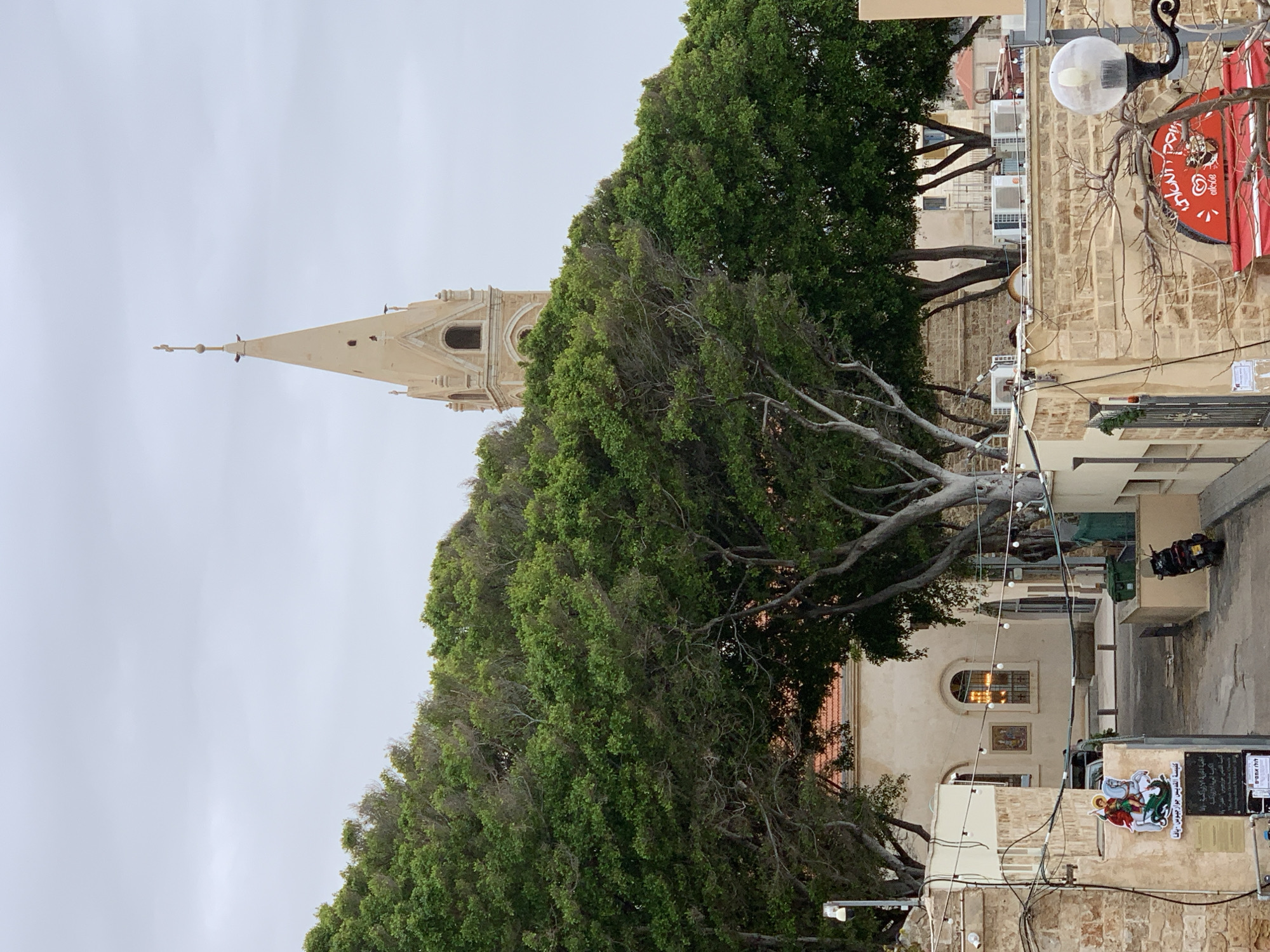 This image depicts an overcast day in Tel-Aviv, Israel, with a view of the Saint George Church in Jaffa. The church's beige spire rises in the background, partially obscured by a large, leafy tree. The surrounding architecture features stone buildings with windows and other architectural details. A red sign with text is visible, as are street lamps and various cables running along the buildings. Signage in Arabic, Hebrew, and English is present on a stone wall, indicating local businesses or informational displays. The overall scene captures an urban setting with a blend of natural and architectural elements.