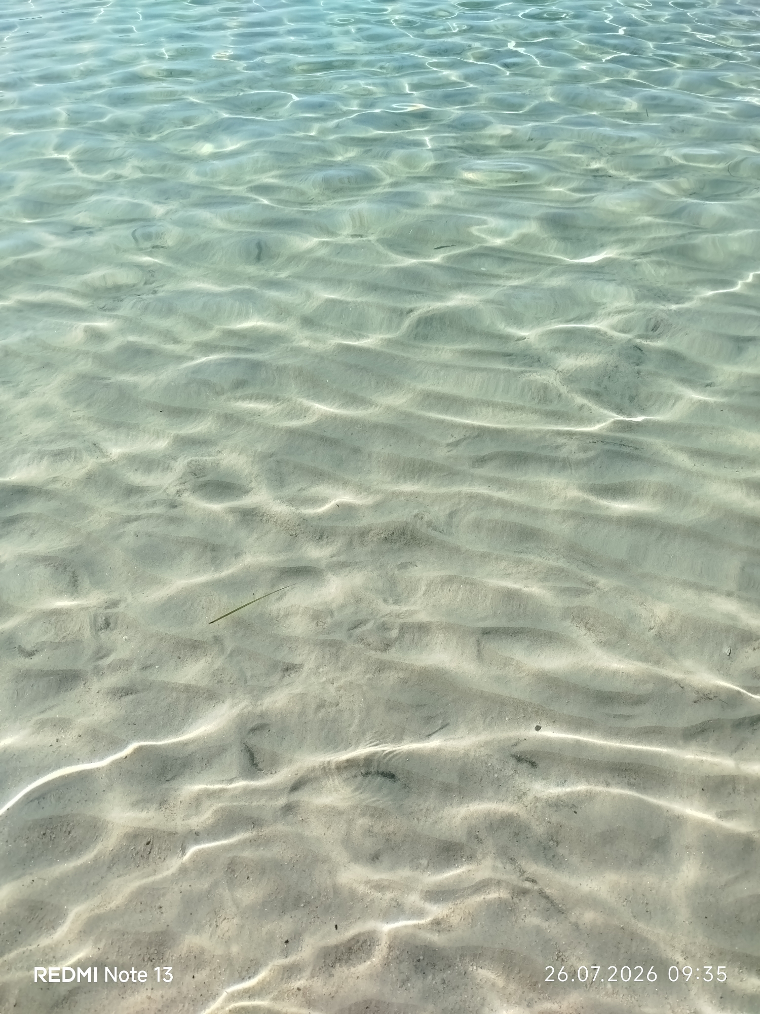 The image shows the shallow sandy bottom of a body of water, likely a beach, with sunlight creating rippling patterns on the sand. The water is clear and has a turquoise hue at the top, transitioning to lighter shades towards the bottom. The sand appears light in color, with some darker specks visible. There is a single green blade of seaweed lying on the seabed in the middle of the frame. No people or animals are visible in the frame. A timestamp in the lower right corner reads "26.07.2026 09:35", and text in the lower left corner reads "REDMI Note 13". The location is identified as Sithonia Municipal Unit, Greece.