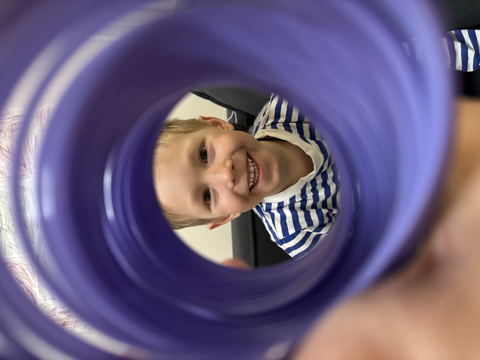 A close-up photograph presents a joyful young child seen through the circular opening of a purple, translucent, ribbed plastic object, likely a tube or bottle. The child, a boy with fair skin and light-colored hair, is positioned centrally within the frame, looking directly at the camera with a wide, beaming smile that reveals his teeth. His bright eyes convey a sense of happiness and engagement. He is dressed in a blue and white striped shirt.
The purple object, which occupies the foreground and frames the subject, exhibits a distinct ribbed texture, creating concentric circles that visually draw attention to the child's face. The unique perspective suggests the photograph was taken from within the object, looking outward.
The background behind the child is softly blurred, indicating an indoor setting. To the left, a light-colored wall with faint patterns, possibly wallpaper or a curtain, is discernible. To the right, a darker, indistinct area suggests furniture or another part of the room. The lighting is bright and even, consistent with an interior environment. No visible text is present in the image. The scene captures a playful moment, reflecting the child's cheerful disposition. This event takes place in Chaikovsky, Russia.