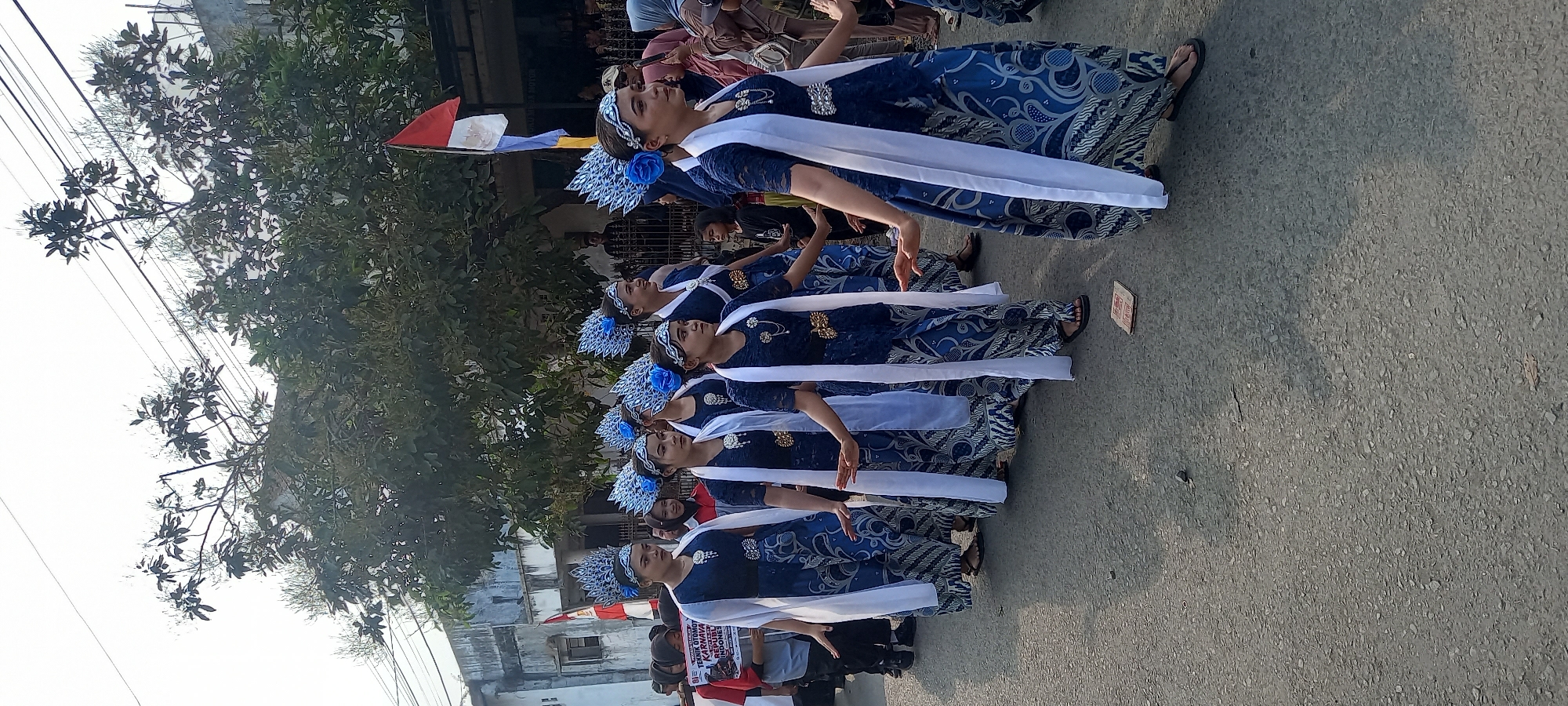 In a street scene in Wanareja, Indonesia, a group of young women in matching traditional blue batik dresses and ornate headpieces participate in a parade. The dresses feature intricate white patterns. They are adorned with white sashes draped over their shoulders and decorated with brooches. Some of the women have elaborate blue feather or flower-like embellishments in their headpieces, alongside silver bands. Their hands are gracefully extended, suggesting they are performing a dance or posing for the procession.
In the background, a red and white Indonesian flag is visible, along with other people spectating the parade. Behind them, buildings and trees line the street. The ground is paved with asphalt. One woman in the foreground holds a sign with red text, which appears to be related to a "Karnaval" or "Republic Indonesia". The overall atmosphere suggests a cultural event or celebration.
