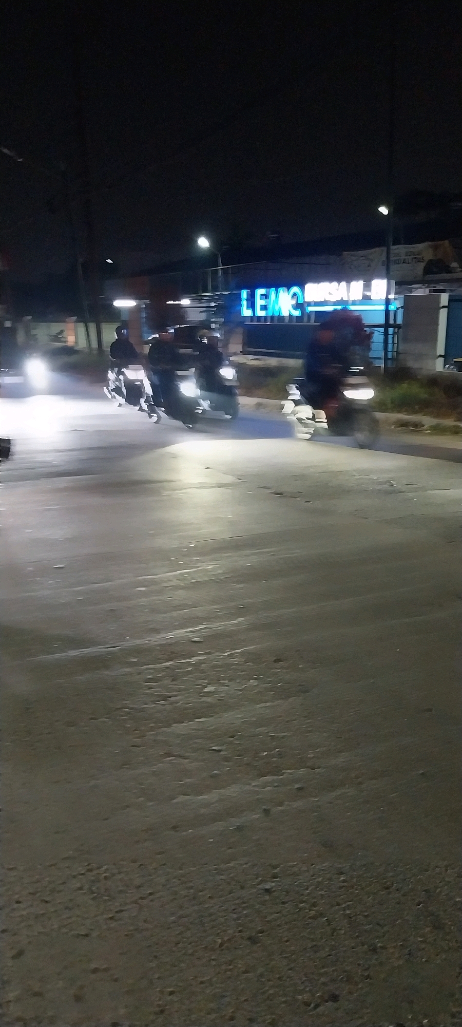 The image captures a nighttime scene on a city street in Kelapa Dua, Indonesia. The foreground is dominated by a dark, wet asphalt road with some gravel and debris. In the middle ground, several motorcycles are in motion, their headlights creating streaks of light. Some of the riders are visible, wearing dark clothing and helmets. Behind the motorcycles, a building with a prominent blue neon sign that reads "LEMO" is visible, along with other illuminated signage and structures. The overall atmosphere is dark and urban, with artificial lighting from the signs and vehicles illuminating the scene. The blur of the moving motorcycles suggests a moderately fast shutter speed was used for the photograph.