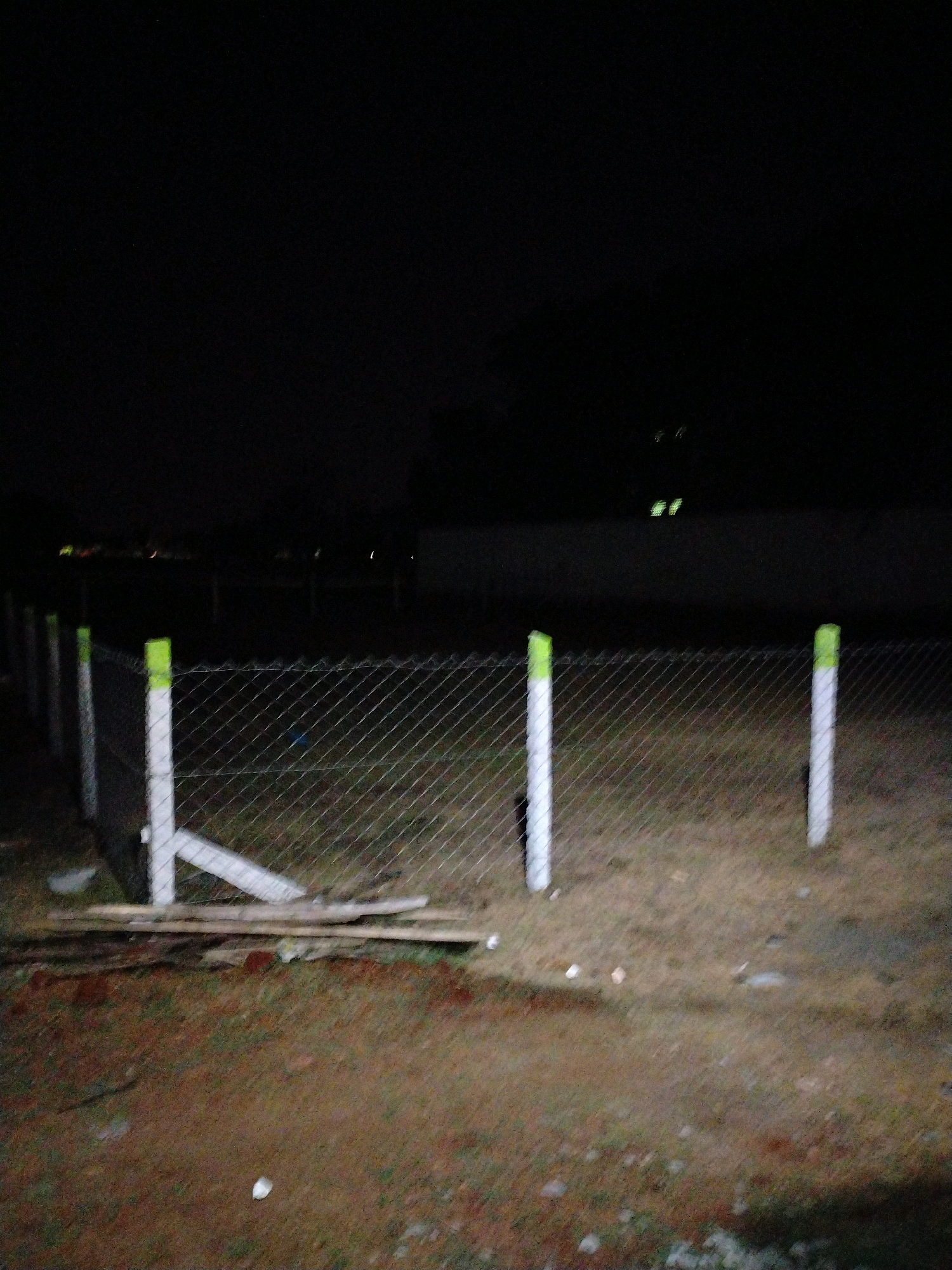 This is a nighttime, low-light image of a chain-link fence with white posts topped with green. The fence is in the foreground and appears to be surrounding an open, grassy area. In the background, there are lights visible in the distance, suggesting it is an urban or semi-urban environment. The ground in front of the fence is a mix of dirt and some sparse vegetation. There is also some discarded wooden material at the base of the fence. The overall scene is dark and somewhat blurry, indicative of a night photograph taken without optimal lighting conditions. This location is within the city of Alwarkuruchi, India.