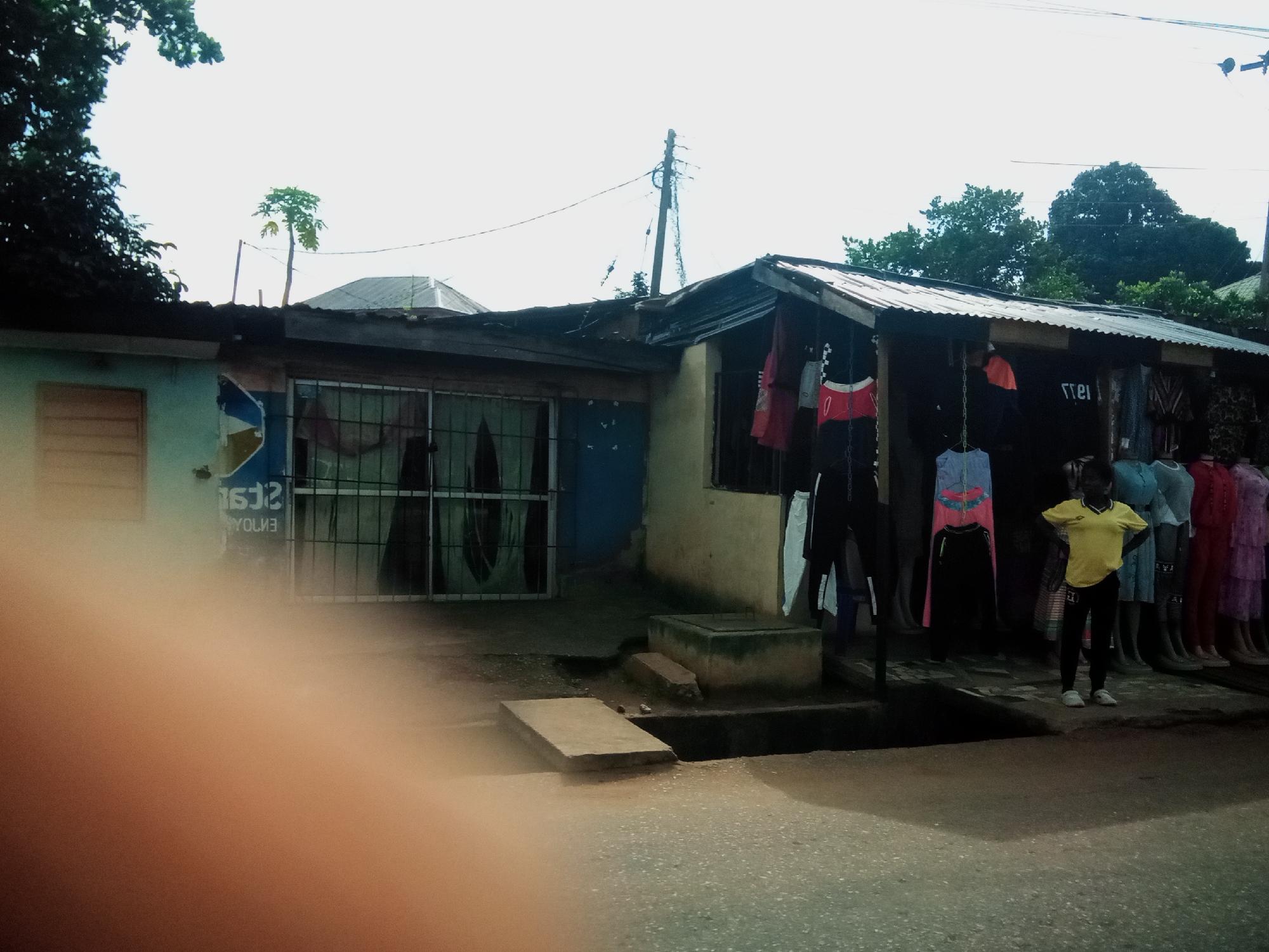 In Nibo, Nigeria, a storefront displays an assortment of clothing for sale. A young individual, possibly a vendor or customer, stands prominently near the clothing racks, wearing a yellow t-shirt and black pants. The storefront itself features a corrugated metal roof and appears to be a simple, open-air structure. Several racks of clothing, including dresses and pants, are visible. In the background, a utility pole with wires stretches across the sky, and trees add to the natural environment surrounding the urban setting. The street in front of the store is paved with asphalt.