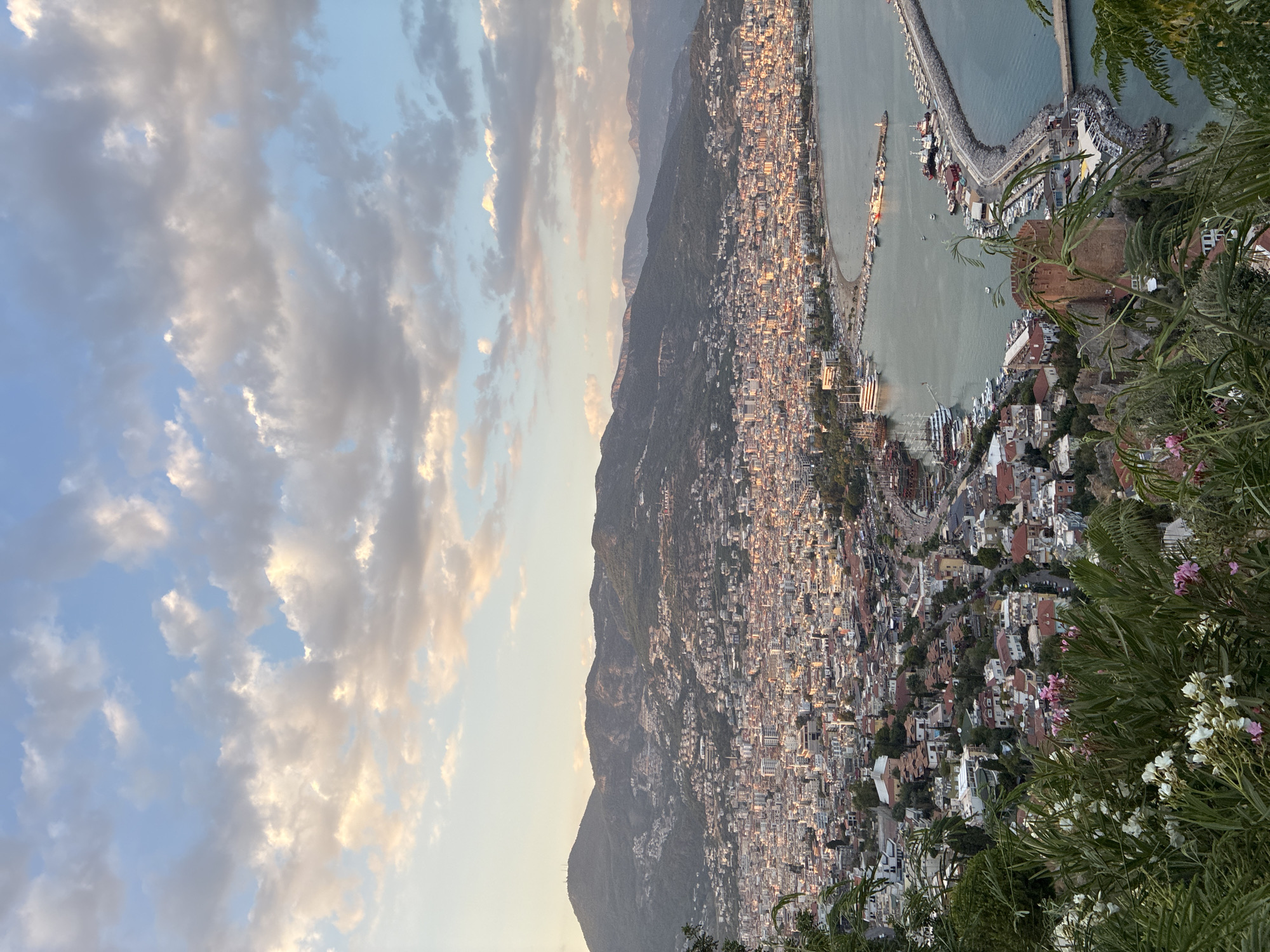 This is a high-angle, eye-level, full shot of the city of Alanya, Turkey, at dusk. The sky is filled with a mix of blue and white clouds tinged with orange and pink hues from the setting sun. Below the sky is a sprawling city with buildings of varying heights and colors, predominantly white and red-tiled roofs, nestled against a backdrop of rolling green hills. The city extends to the coast, where a harbor is visible, filled with numerous boats and yachts. A prominent stone fortress with a red-brown tower stands on a hill overlooking the harbor and the city. Lush green foliage, including flowering plants, is in the foreground, framing the panoramic view of the city. The lighting suggests a warm, late afternoon or early evening atmosphere, with shadows beginning to lengthen across the urban landscape.