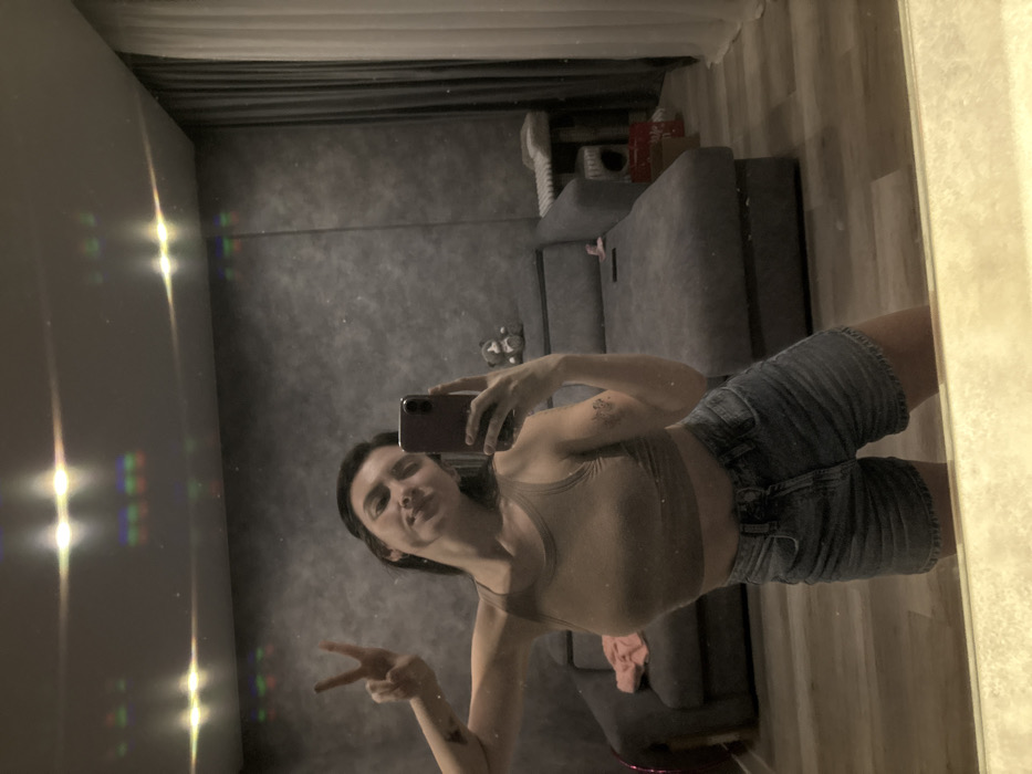 An indoor mirror selfie captures a young woman posing in what appears to be a living room or bedroom. The image is oriented with the subject lying horizontally, indicating it was likely taken with the phone held vertically and then the photo rotated for viewing.
The woman, with dark hair, is visible from approximately the chest down to the mid-thigh. She is wearing a fitted light brown or beige athletic crop top and light blue denim shorts. Her left arm is extended towards the camera, holding a dark-colored smartphone, possibly purple or dark red, with two visible camera lenses. Her right arm is bent at the elbow, and her hand is held up, displaying a "peace" sign (V-sign) with her index and middle fingers extended. She has a slight smile on her face. A small, dark tattoo or mark is visible on her left upper arm.
The background, visible through the mirror's reflection, shows a room with a somewhat dim and warm ambiance. On the left side of the reflection, bright, multi-colored light flares and streaks are prominent, suggesting reflections from a light source or window, possibly creating diffraction effects due to the mirror surface or camera lens. Beyond the subject, in the reflected space, there is a dark grey, textured wall and a grey sofa or couch with a subtle pattern. On or near the sofa, various items can be vaguely discerned, including what looks like a pair of white sneakers and possibly a light-colored blanket or clothing item. The floor, visible at the bottom of the reflected image (right side of the actual room), is made of light-colored wooden planks. The mirror surface itself appears to have some dust or minor imperfections.