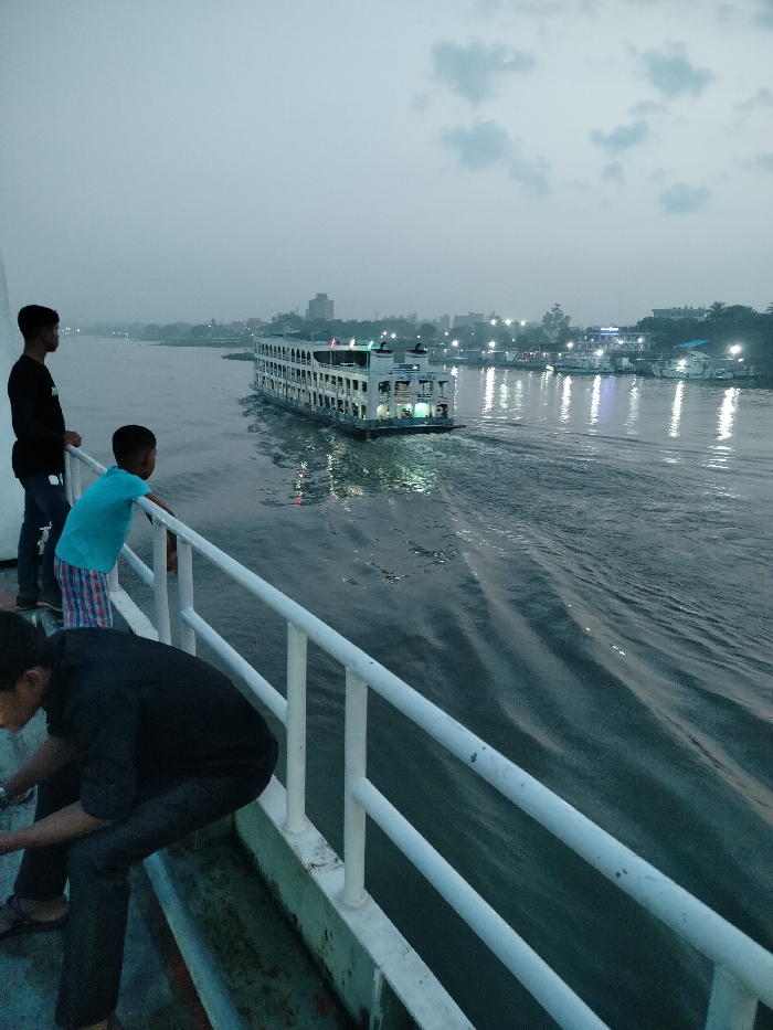 Three people stand on a boat in Zazira, Bangladesh, facing a large, white, multi-level passenger ferry traveling away from them across the river. The boat is moving, as evidenced by the rippling water. The sky is gray and cloudy. The ferry is lit by the setting sun. The people on the boat are watching the ferry pass by. Two of the people are young boys wearing short-sleeved shirts and pants. The third person is an adult man wearing a black shirt and dark pants. He is leaning on the white railing of the boat. The boat is in the foreground. The ferry is in the middle ground. The cityscape is in the background. The river is large and the water is calm and smooth. The sky is overcast. There is a slight mist in the air. The ferry is illuminated by the setting sun, casting a warm glow on the water. The people on the boat are silhouetted against the setting sun. The scene is peaceful and serene. The coordinates provided are: (latitude: 23.654, longitude: 90.459).