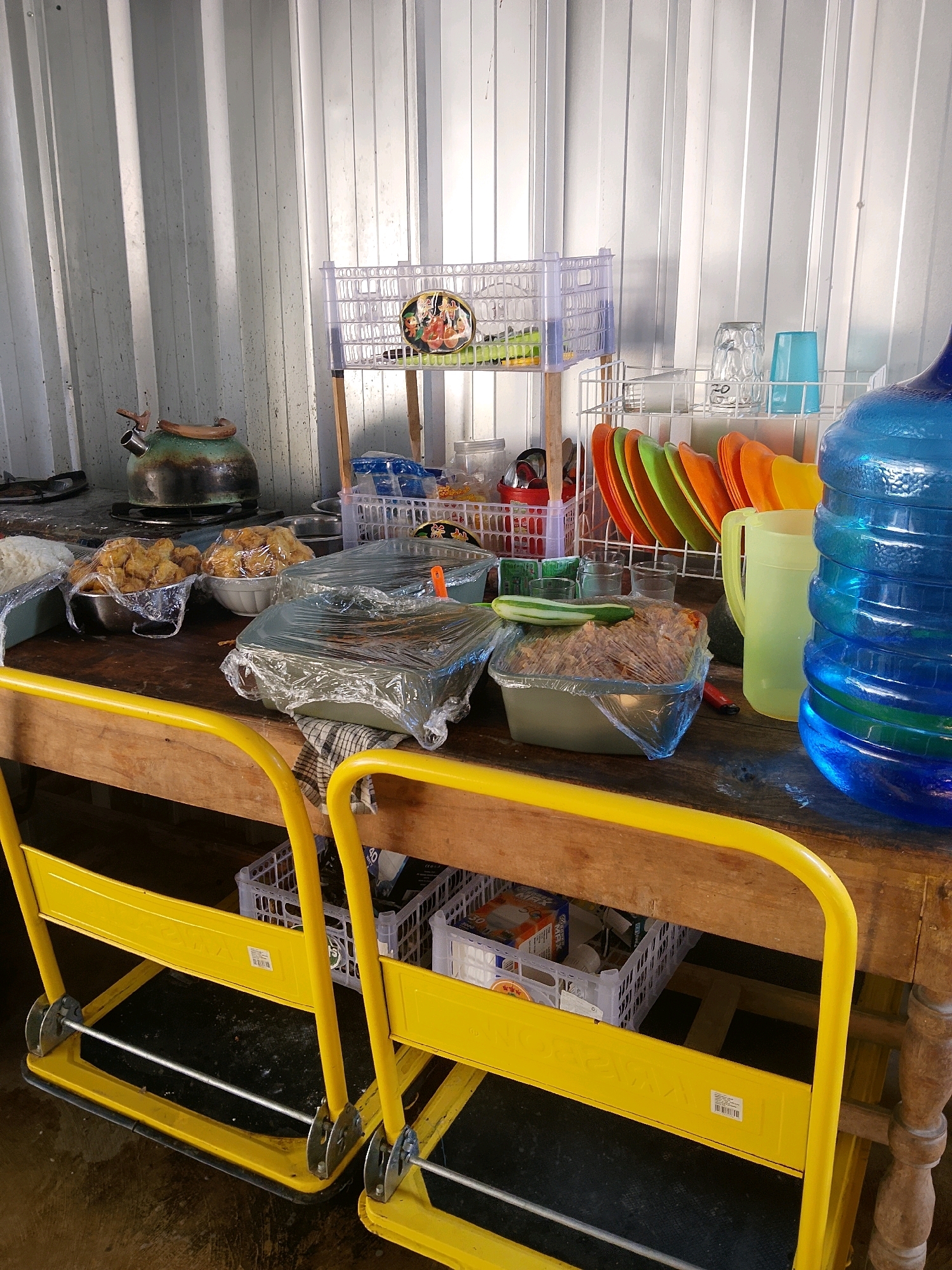 This is a medium shot, eye-level, outdoor, daylight photograph taken in Negeri Agung, Indonesia. The scene depicts a rustic wooden table laden with food and kitchenware, set against a backdrop of corrugated metal walls. In the foreground, two bright yellow utility carts are partially visible, their metal handles curved.
The table is covered with various dishes, many of them wrapped in clear plastic. To the left, a metal kettle with a brown lid rests on what appears to be a stovetop, suggesting a food preparation or serving area. Bowls filled with fried food items, also covered in plastic, are placed centrally. Further to the right, a large, clear plastic container holds a dish topped with a slice of cucumber, and another container is filled with a meat dish, both wrapped in plastic.
Behind the table, a white wire rack holds stacks of colorful plates in orange, green, and yellow, as well as several glasses, including a clear glass with visible writing and a light blue plastic cup. A yellow pitcher is also present on the rack. To the far right, a large, blue water dispenser dominates the frame, its curved surface reflecting the light.
On a higher shelf, a white plastic basket contains a round decorative item featuring figures, possibly religious or cultural in nature. Additional white plastic baskets filled with various items, including boxes, are visible beneath the table. The overall impression is of a busy, functional outdoor food service area.