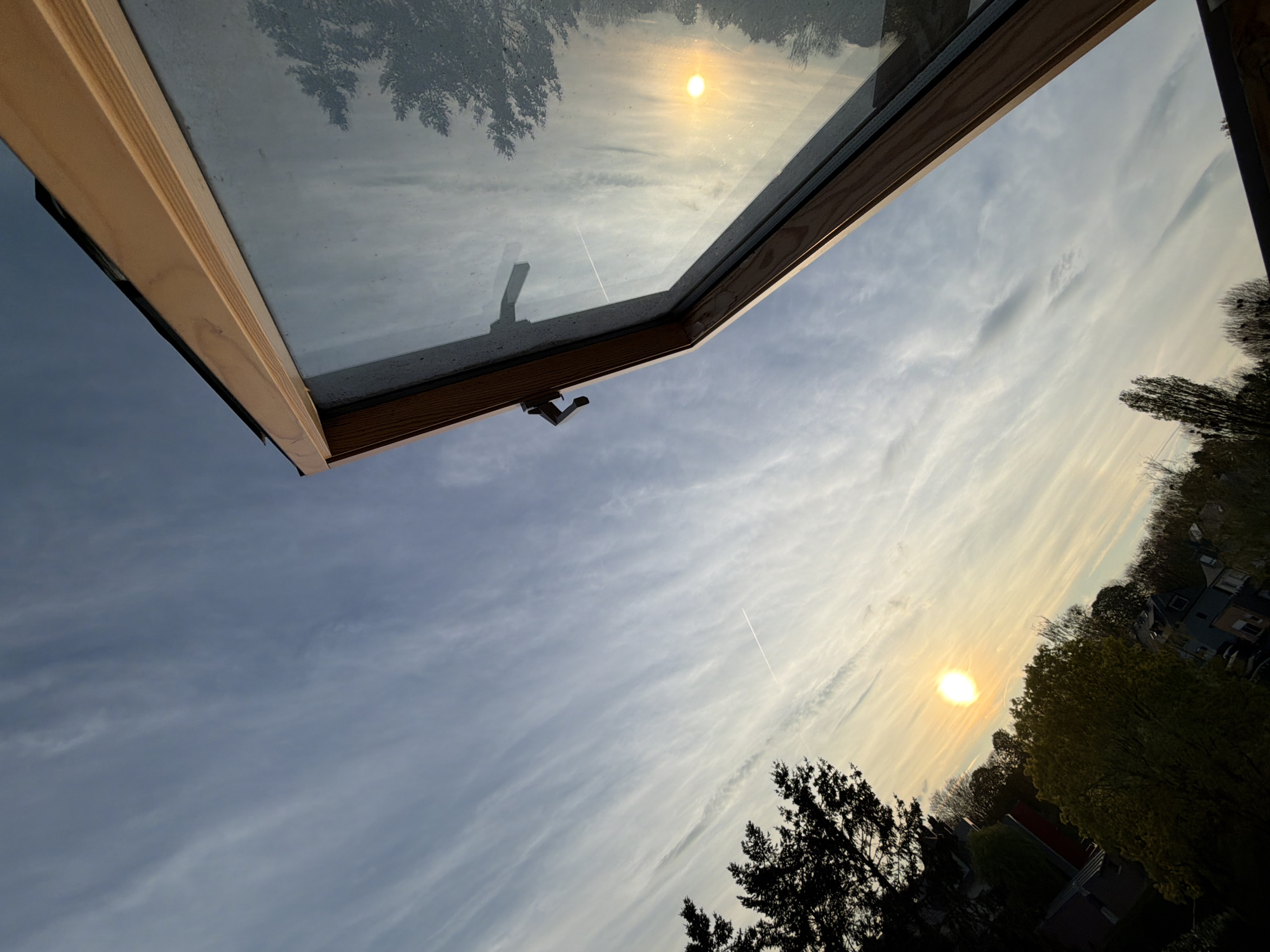 The image shows an upward-angled view through an open skylight. The wooden frame of the skylight is visible, with the glass partially reflecting the sky and trees. The sky is filled with wispy clouds and a bright sun, reflecting off the glass. Outside the window is a view of rooftops and trees, typical for Esch-sur-Alzette, Luxembourg. A contrail from an airplane is also visible in the sky. No people are visible.