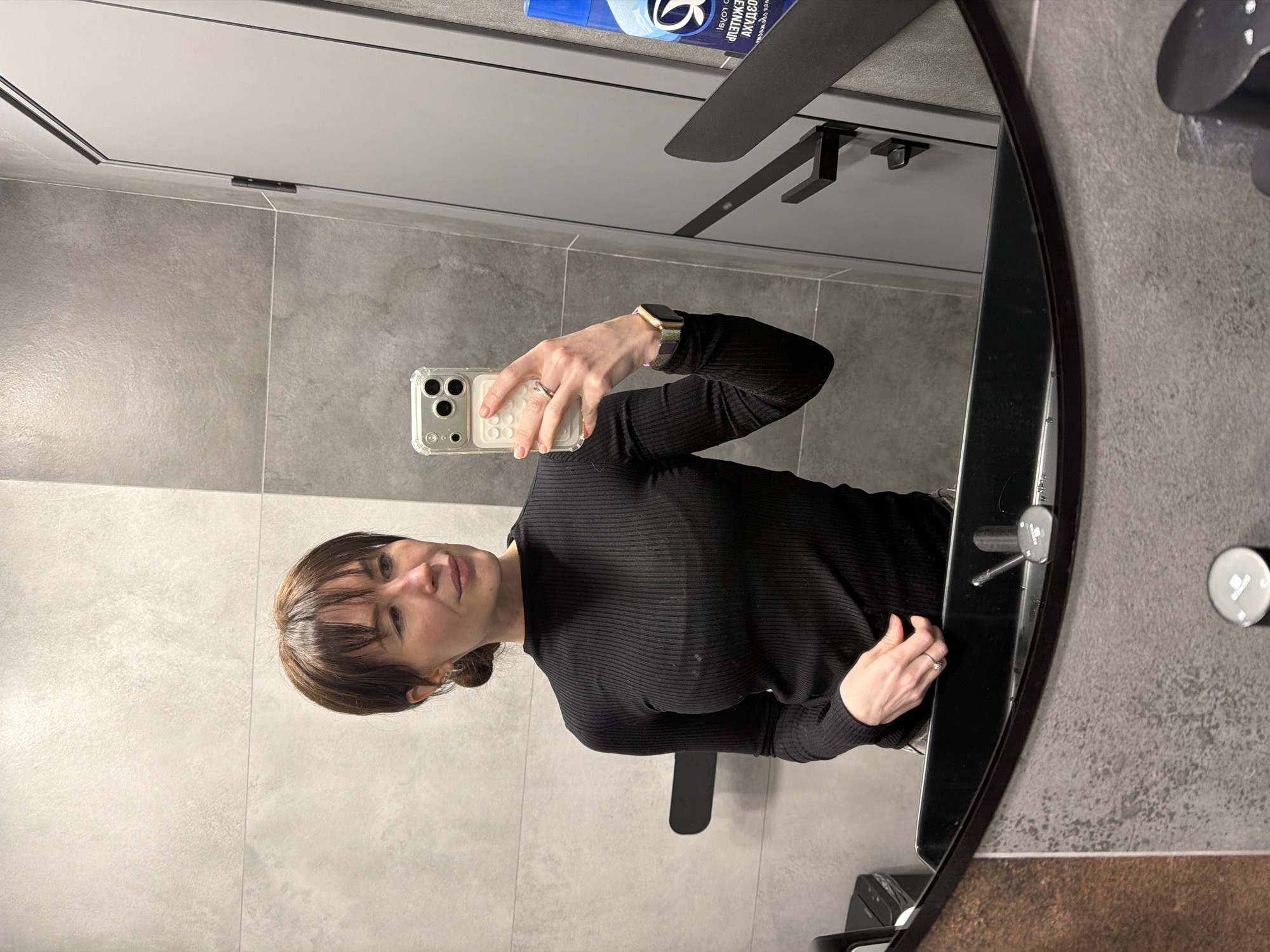 A woman is captured in a mirror selfie, holding a smartphone with a clear case in her right hand. She has brown hair with bangs, styled away from her face, and is wearing a black ribbed long-sleeved top. On her left wrist, a gold-toned smartwatch is visible, and she wears a silver ring on her left ring finger. She has a neutral expression with a slight smile.
The setting appears to be a modern public restroom, characterized by light grey, textured wall tiles. The mirror itself has a dark, slightly curved frame on the right side. In the reflection, above the woman's head, a blue bottle with Cyrillic text is clearly visible. The text includes "ЛЕВЕНЯТ АКЦИОН" and "ЖИДКОЕ," which translates to "LIQUID," indicating a soap or cleaning product. Other reflective elements include a white ceiling and a sleek black fixture, possibly a shelf or part of a ventilation system, above the bottle. Below the woman's right arm, another black fixture, resembling a tap or dispenser, is reflected. The environment is well-lit and clean, consistent with a commercial or institutional building in Perm, Russia.