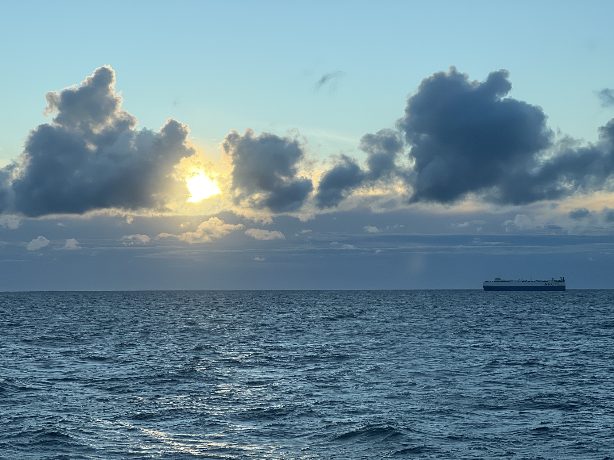 An expansive view of the sea under a sky filled with dramatic clouds at either sunrise or sunset. The sun is breaking through a cluster of dark clouds on the left, casting a bright, diffused light. The sea is choppy with visible ripples and waves, reflecting the muted colors of the sky. On the right horizon, a large cargo ship is sailing, appearing small against the vastness of the ocean and sky. The scene conveys a sense of solitude and the grandeur of nature. The location is identified as an unspecified coastal area in Poland, though the specific city is unknown.