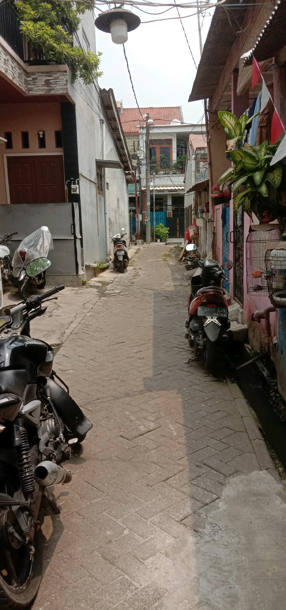 The image depicts a narrow, paved alleyway in a residential area of Tangerang, Indonesia. The alley is lined with buildings on both sides, featuring a mix of architectural styles. Several motorcycles are parked along the sides of the alley, indicating their common use for transportation in the area. One motorcycle on the left is partially covered with a clear plastic sheet. The alley is paved with brick pavers and shows signs of age and wear. There are also electrical wires and a light fixture suspended overhead. In the background, more buildings and rooftops are visible, suggesting a dense urban environment. The scene is illuminated by natural daylight, casting shadows on the alleyway. No people are visible in the image.