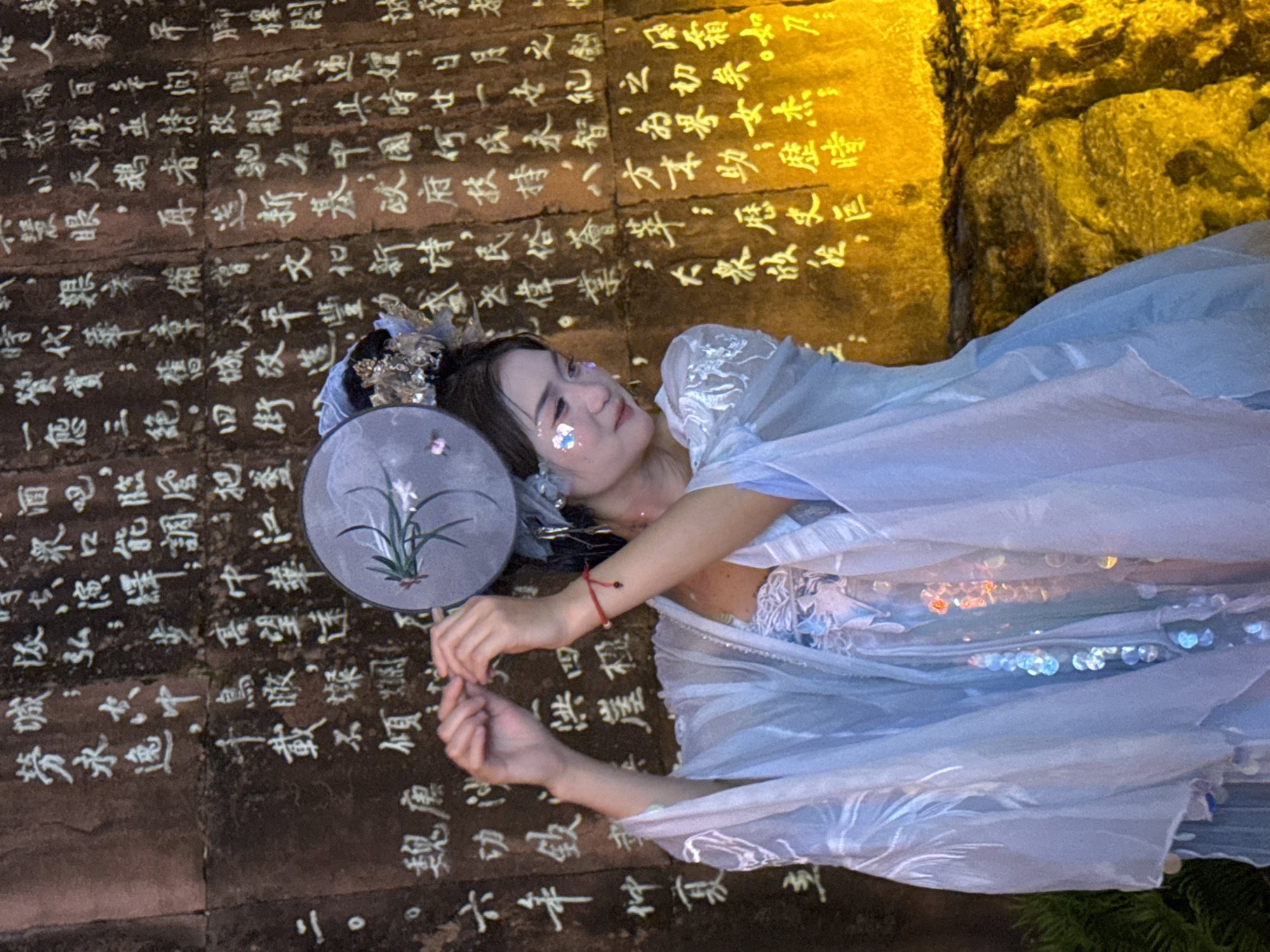 The image shows a young woman dressed in a light blue, flowing traditional Chinese costume, possibly Hanfu, with ornate silver embroidery. She is adorned with sparkling glitter on her face and a delicate headdress. She is holding a round, grey fan with a painted floral design. The woman is standing in front of a large stone wall inscribed with vertical columns of white Chinese calligraphy. The stone appears weathered and uneven. The wall is illuminated by a warm, yellowish light, casting shadows and highlighting the texture of the stone and the inscriptions. The background also shows hints of green foliage on the left. The overall scene evokes a sense of historical or cultural significance, possibly a performance or a photoshoot in a location like the Yuzhong District of Chongqing, China, which is known for its historical sites and cultural heritage.