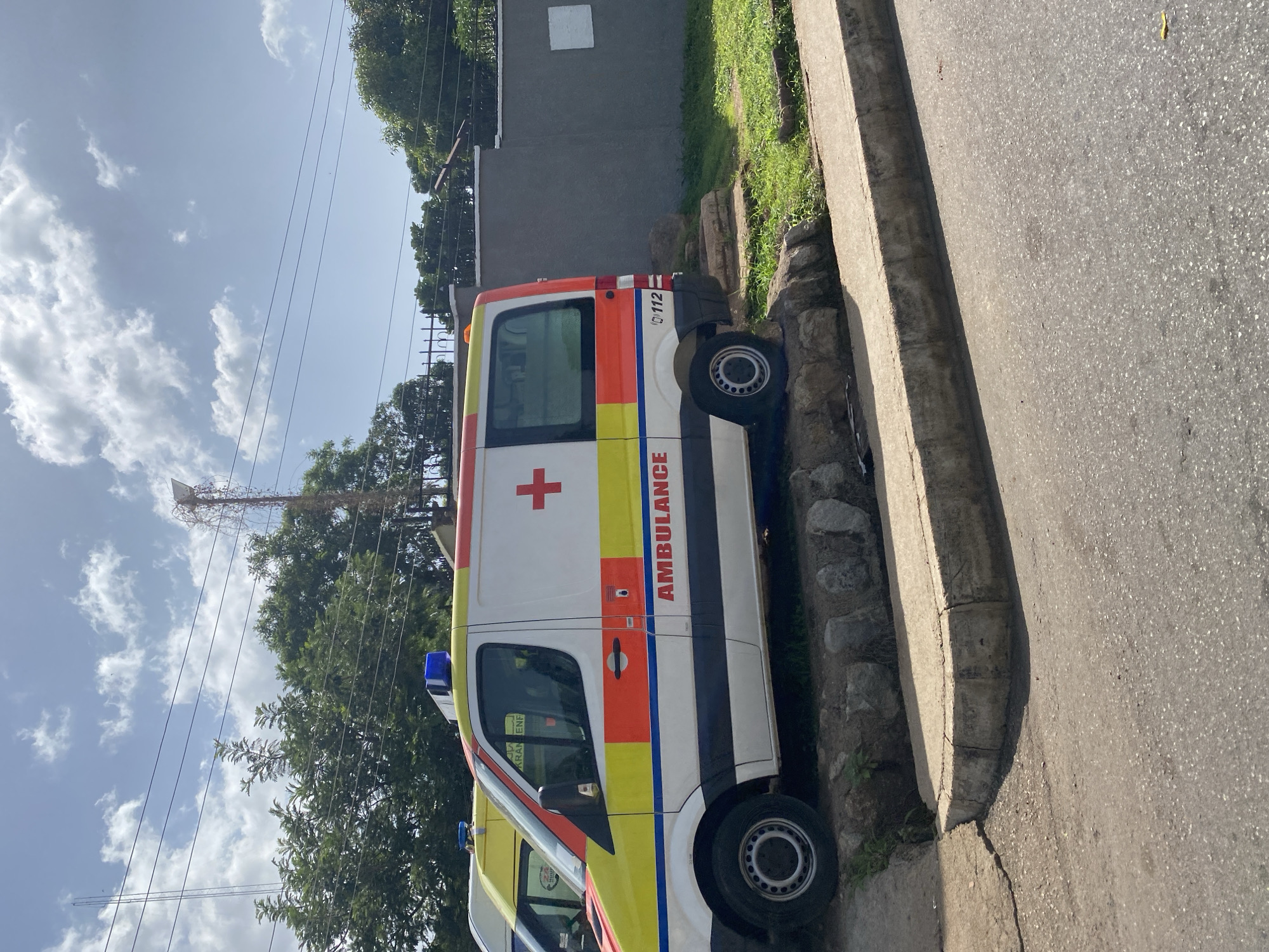 The image shows a white ambulance with red and yellow markings parked on a slight incline next to a grey wall. The ambulance has "AMBULANCE" written in red on its side and a red cross symbol. There is another yellow vehicle partially visible in the background. The scene is outdoors, with green trees and a blue sky with white clouds. A power pole with wires is visible behind the trees. The ambulance is parked on a dirt and stone surface next to a concrete curb and a paved road. The environment suggests an urban setting, possibly in a medical facility or residential area in Jos, Nigeria.