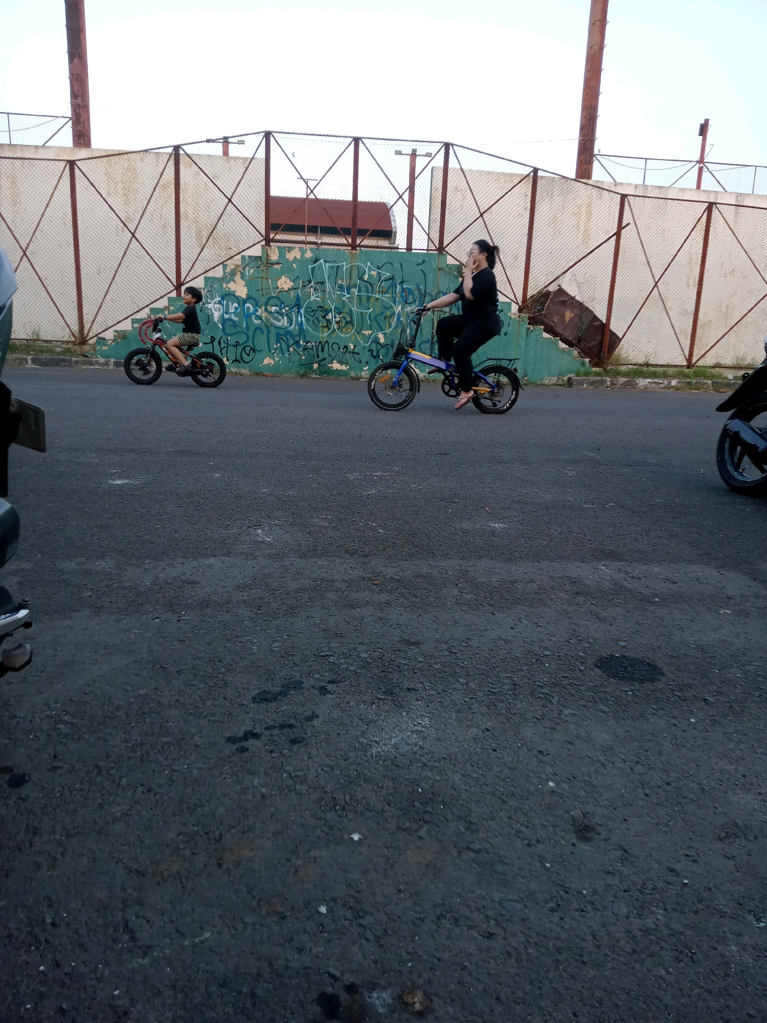 An outdoor, medium-range, eye-level shot captures a boy on a red bicycle and a woman on a blue bicycle riding on an asphalt road. The boy, facing left, is wearing a black t-shirt and striped shorts. The woman, facing right, is wearing a black t-shirt and black pants, with her right hand on her chin. Behind them, a green wall is covered in graffiti, and a chain-link fence runs along the top of the wall. A large, rusty metal pole stands to the left of the fence. The background shows a clear sky and a building with a brown roof. In the foreground, parts of a motorcycle are visible on the left and right sides of the frame, indicating this scene might be in a parking lot or a street where motorcycles are common. The overall impression is a casual outdoor activity, possibly in a urban or semi-urban environment in Karawang, Indonesia, given the presence of graffiti and the style of fencing and buildings often seen in that region.