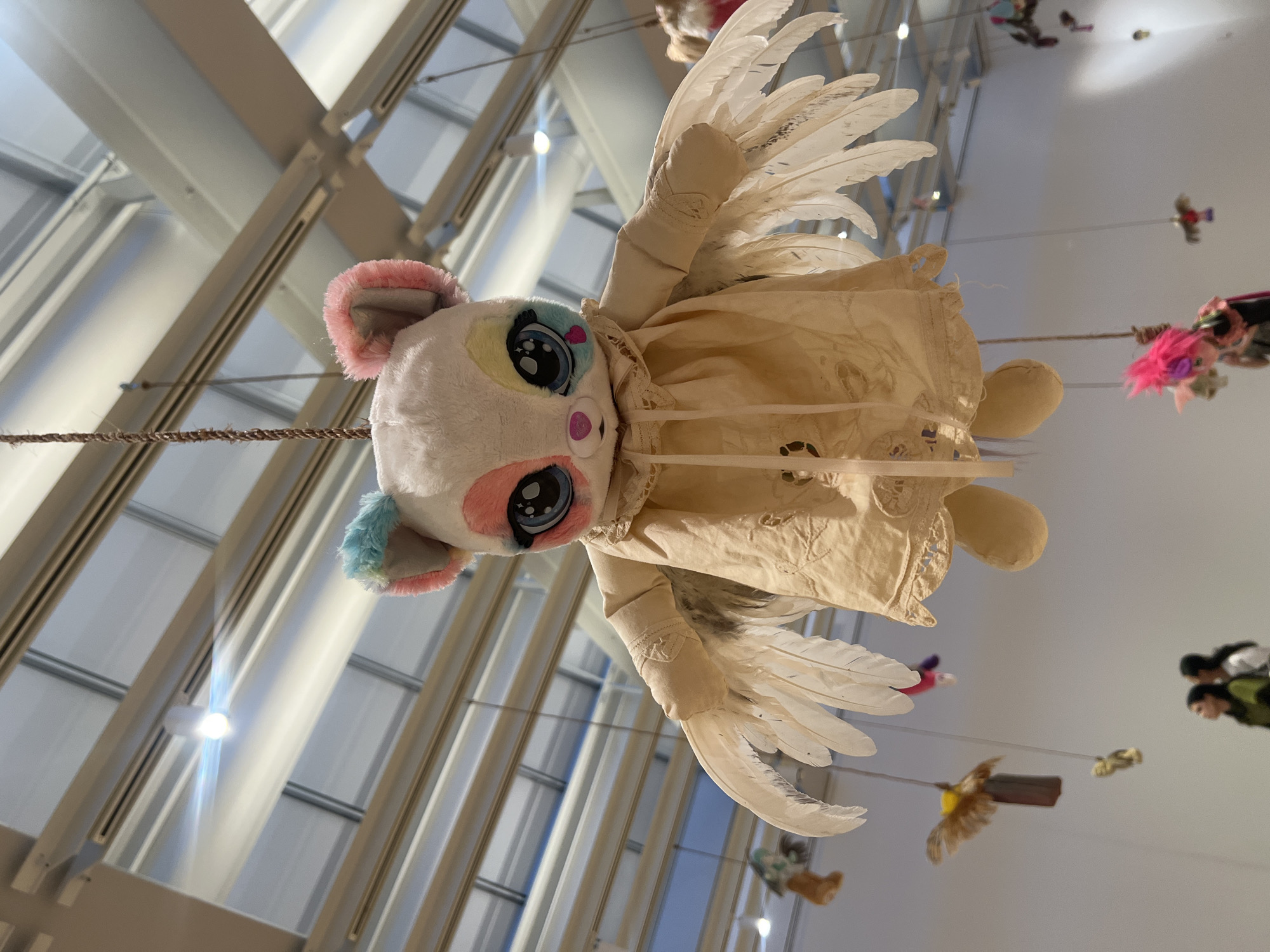 A photograph shows a stuffed animal with wings and a dress suspended in the air in front of a large window. The stuffed animal has a white head with pink and blue fur on its ears, and large, cartoonish eyes. Its body is covered by a cream-colored dress with lace detailing. Two wings made of white feathers are attached to the back of the dress. The animal appears to be floating due to the way it hangs from the ceiling. The large window provides a view of a modern-looking building with a steel and glass facade. Other objects are hanging in the background. Given the unique nature of the installation and the architectural style of the building, it is likely that the photograph was taken inside an art gallery or museum in New York City.