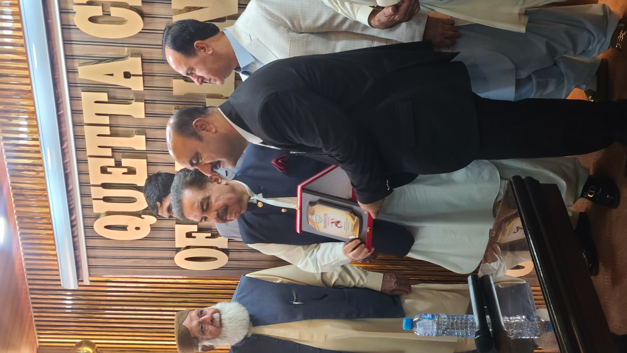The photograph captures a group of men gathered in what appears to be an indoor event in Quetta City Tehsil, Pakistan. One man is holding a framed award. Behind them is a wall with large, gold letters spelling "QUETTA CHAMBER." The men are dressed in a mix of formal and traditional Pakistani attire. There are microphones and water bottles visible in the foreground. The environment suggests a formal or semi-formal gathering, possibly an awards ceremony or an inauguration event.