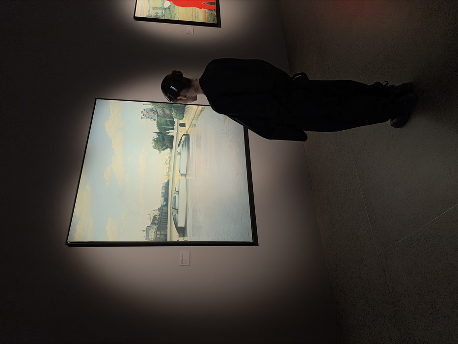 A person wearing all black faces a large painting of a cityscape. The painting dominates the left side of the image, bathed in light. Above it, a smaller painting with red hues is visible. The person stands to the right, their form outlined against the grey tiled floor and walls. The overall ambiance is dim, typical of an art gallery setting.