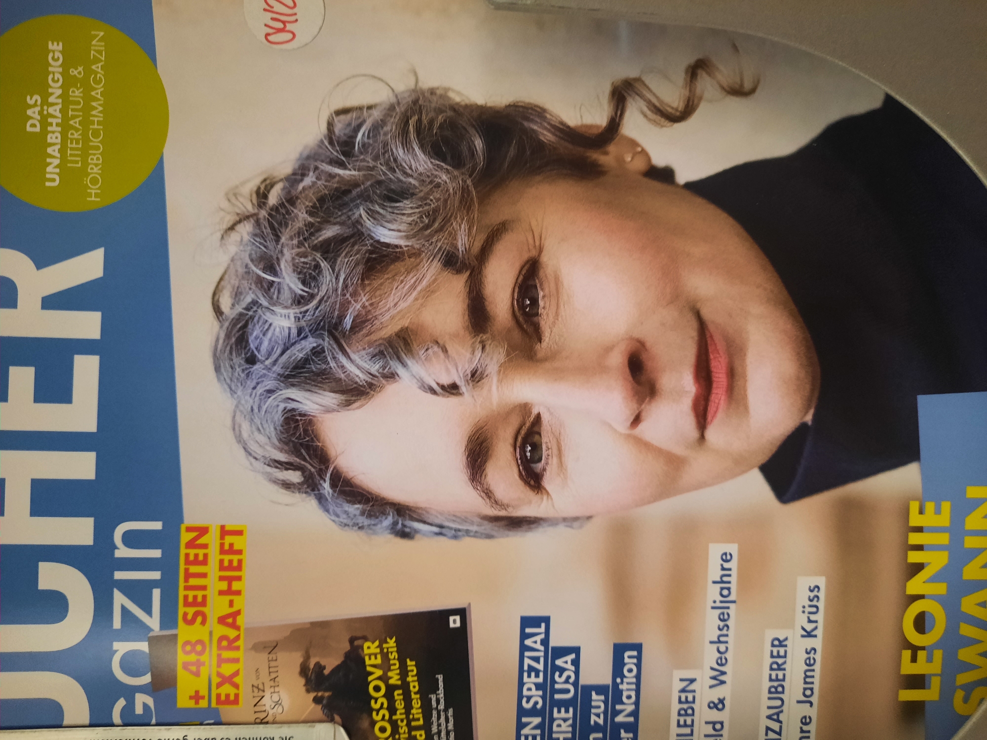 A close-up, eye-level, portrait-oriented photograph captures a portion of the cover of a German magazine. The magazine appears to be a literary and audiobook publication, as indicated by the text "DAS UNABHÄNGIGE LITERATUR- & HÖRBUCHMAGAZIN" (The Independent Literature & Audiobook Magazine) in a lime green circle on the upper right of the cover. Below this, the word "gazın" is visible in large blue font, suggesting the magazine's name. A yellow strip prominently displays "+ 48 SEITEN EXTRA-HEFT" (48 Pages Extra Insert).
The dominant subject is the right half of a woman's face. She has fair skin, with subtle makeup highlighting her cheeks and lips. Her eyes are a light brown, and she has expressive eyebrows and fine wrinkles around her eyes and mouth. Her hair is a striking blend of grey and darker strands, styled in loose, curly tendrils that frame her face. She is wearing a dark, high-necked garment, possibly a turtleneck sweater.
To the left of the woman's face, several text blocks are visible. One advertises a "CROSSOVER Zwischen Musik und Literatur" (Crossover Between Music and Literature). Further down, text in blue boxes reads "EN SPEZIAL IHRE USA" (EN SPECIAL YOUR USA), "rn zur er Nation" (rn to the Nation), "RZAUBERER" (RZAUBERER - likely a title), and "hre James Krüss" (hre James Krüss). At the bottom left, large yellow letters spell out "LEONIE SWANN".
A red handwritten or stamped date, "04/12", is present on the right side of the magazine cover, near the woman's hair. The background is a muted, slightly textured beige. The image does not contain any specific geographical markers or identifiable landmarks that would allow for its location to be determined within the city of Rosenheim, Germany. The scene depicts a static magazine cover.