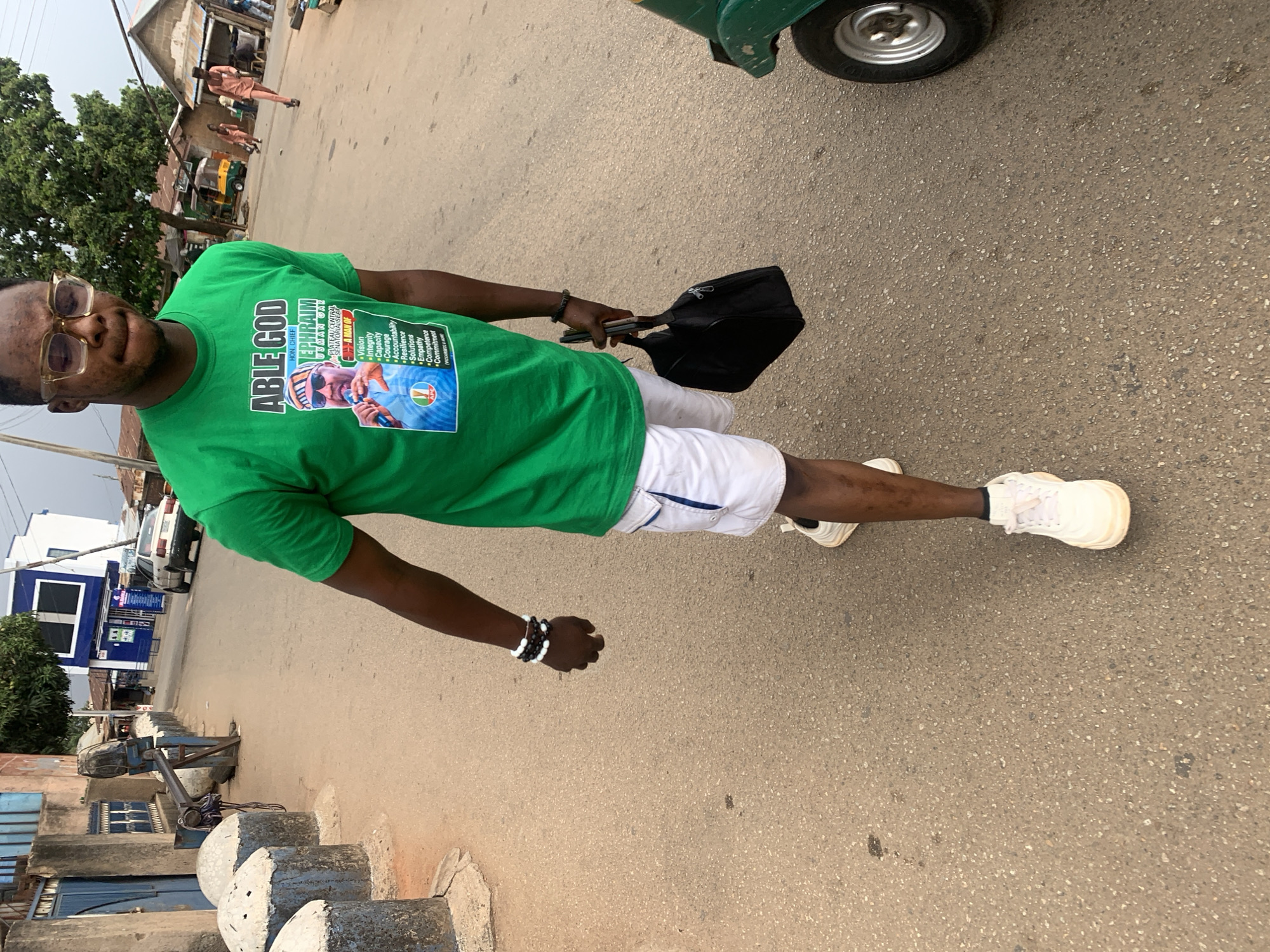A man wearing glasses, a green t-shirt with political text, white shorts, and white sneakers stands on a paved road. He holds a black bag in his right hand and wears bracelets on his left wrist. The road is lined with low concrete barriers, and a building with blue window frames is visible in the background. The T-shirt reads "ABLE GOD, HON. CHIEF EPHRAIM USMAN CAR, PLATEAU CENTRAL SENATORIAL SEAT, A MAN OF," followed by words like "Vision, Integrity, Capacity, Courage, Accountability." In the background, there are a few people and vehicles including a three-wheeled vehicle. The location appears to be in Jos, Nigeria, based on the political context of the shirt and the general environment.