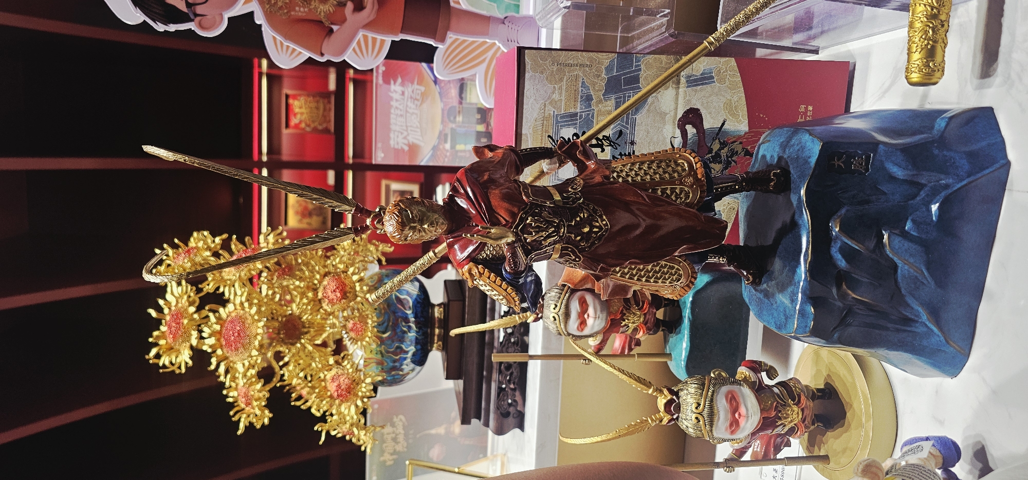 This is a medium shot, eye-level photograph of a display featuring several Monkey King figurines and a golden floral arrangement. The figurines are intricately detailed, with the central figure being the largest and most prominent, adorned in red and gold armor, holding a golden staff. Two smaller Monkey King figurines are positioned in front and to the left of the main one. Behind the figurines, a large golden floral arrangement in a blue and white patterned vase is visible. The background suggests an indoor setting, possibly a store or exhibition, with shelves displaying more items and red-colored accents. The lighting is soft and highlights the metallic textures of the figurines and the floral arrangement. The photograph appears to be taken in a city, however, the provided context does not specify the city or district, thus an exact location cannot be determined.