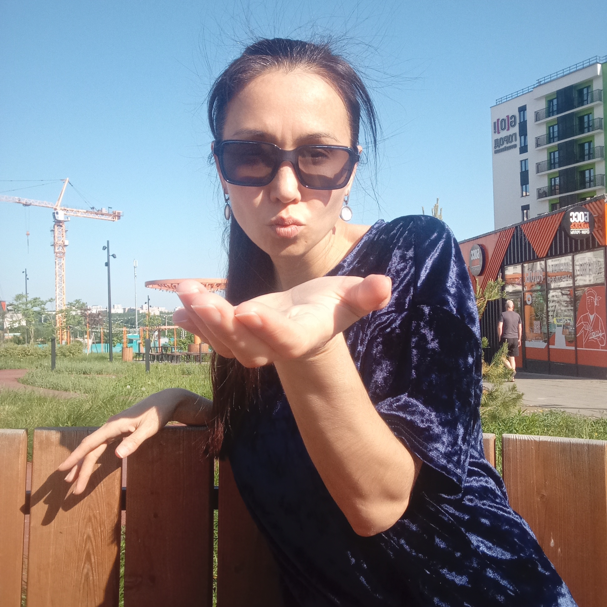 A woman with dark hair pulled back into a ponytail, wearing dark sunglasses and a dark blue velvet top, is sitting on a wooden bench outdoors. She is extending her right hand as if blowing a kiss. In the background, there is a grassy area, a construction crane, and a modern building with Cyrillic text that includes the name "DOROGOY," and another building with the number "3303" on the side. A man in a gray shirt and dark shorts is walking near the 3303 building. The photograph appears to be taken in a park-like setting in Pervomaysky, Russia, based on the Cyrillic text on the buildings and the overall aesthetic.