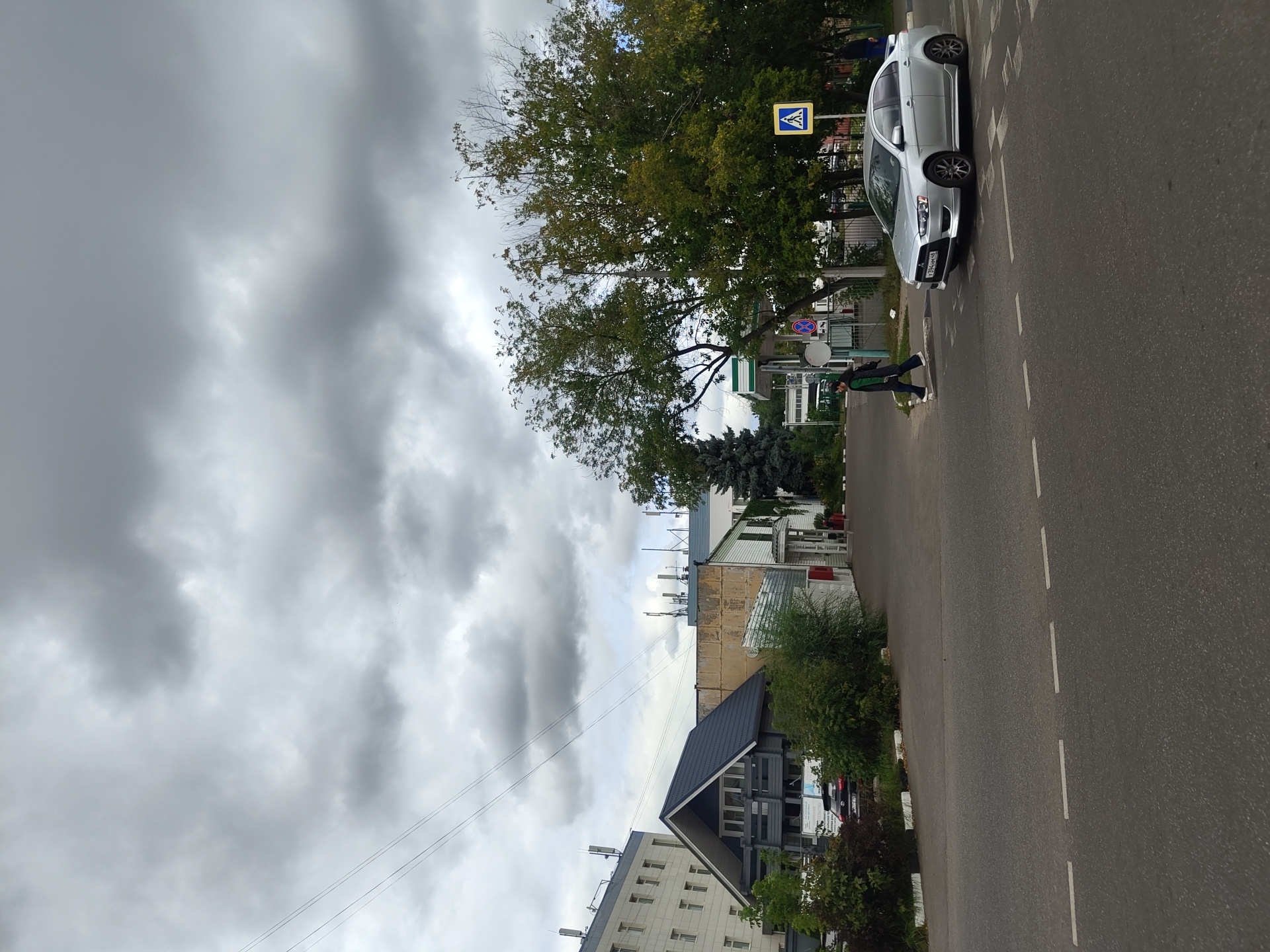 The image depicts a street scene in Zhukovsky, Russia, under a heavily overcast sky. In the foreground, a paved road with dashed white lane markings is visible. A silver sedan is crossing a pedestrian crossing from right to left. A person wearing a dark jacket and pants is walking across the pedestrian crossing from left to right, in front of the car. To the right of the pedestrian crossing, a yellow square sign with a blue border and a white silhouette of a pedestrian indicates a crosswalk.
In the middle ground, there is a sidewalk and a gas station with a green and white canopy. Behind the gas station, several trees with green and yellowing foliage are present, with one large evergreen tree to the left. Several buildings are visible in the background. To the left, a multi-story light-colored building with numerous windows and what appear to be cellular antennas on its roof stands out. Adjacent to it is a smaller, modern-looking house with a dark pitched roof and balconies. Further to the right, other buildings with a more industrial or commercial appearance are partially obscured by trees. Power lines are strung across the upper portion of the image. The overall atmosphere is muted due to the cloudy weather.