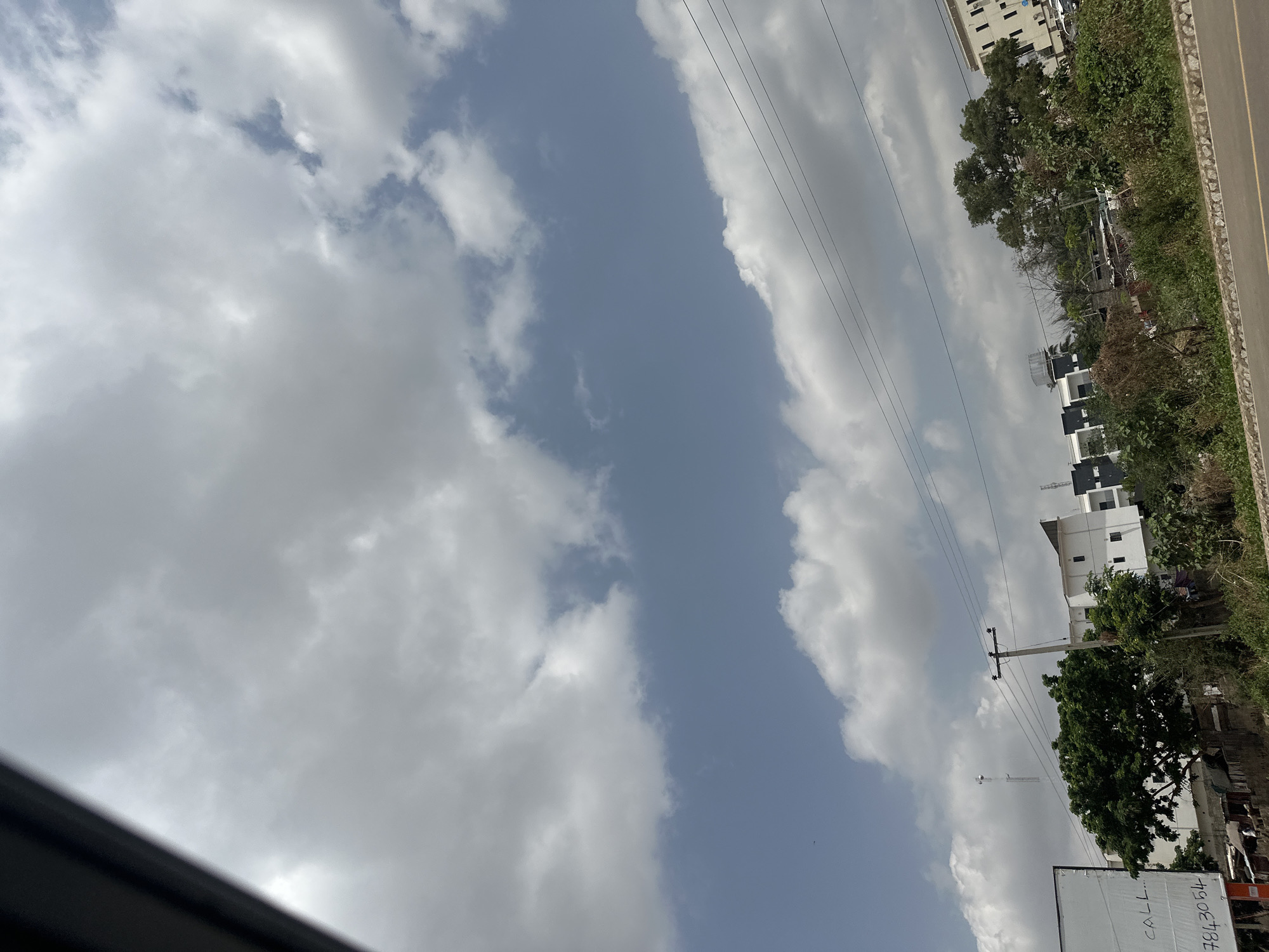 This media file presents a daytime view of a semi-urban or suburban landscape in Abuja, Nigeria, characterized by a mix of natural elements and human infrastructure. The image is oriented vertically, showing the sky on the left and the ground on the right (when rotated 90 degrees clockwise to a standard landscape orientation).
The dominant feature is the sky, which occupies a large portion of the frame. It displays a bright blue color interspersed with numerous large, white, and grey cumulus clouds, indicating fair weather. Patches of clear blue sky are visible between the cloud formations.
On the right side of the image, a grey road with a distinct yellow dividing line runs along the edge. Adjacent to the road, there is a dense strip of green vegetation comprising various trees and shrubs. Several multi-story buildings, some painted white and others featuring darker facades, are visible peeking through and above the trees. Utility poles supporting multiple power lines extend diagonally across the sky and landscape. In the distance, a tall, slender tower, possibly a communication or cell tower, rises above the buildings and trees.
In the lower right corner, a white rectangular billboard or sign is partially visible. On this sign, the word "CALL..." and the numerical sequence "7843054" are distinctly legible, likely indicating contact information. No people or specific activities are observable in the scene, which appears to be a static capture of a street view in daylight.