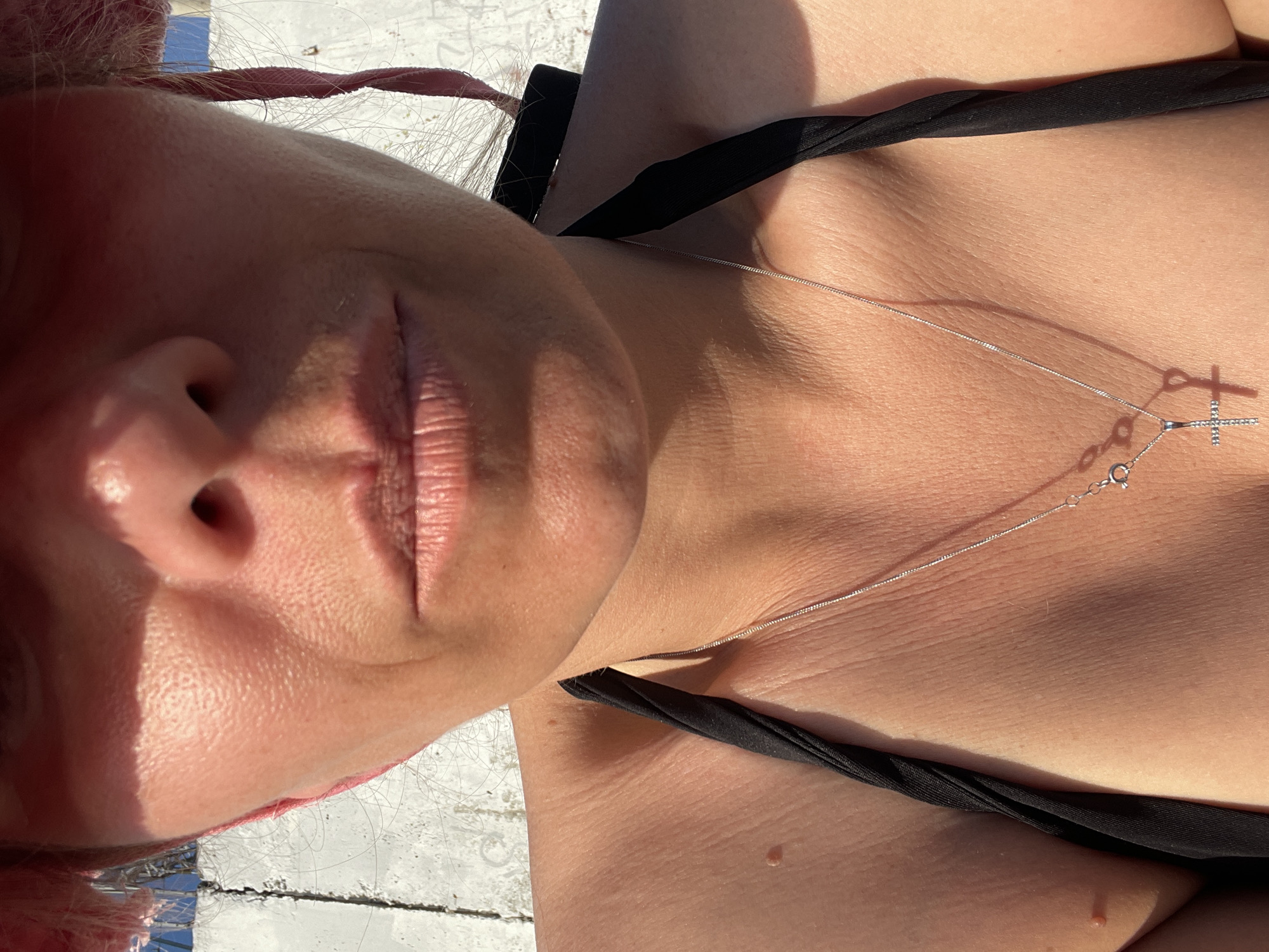 This is a close-up, portrait-oriented photo taken from a low angle, featuring a person's face and upper chest. The person appears to be lying down, possibly outdoors, as indicated by the bright sunlight casting shadows on their skin. They are wearing a black bikini top, and a thin silver necklace with a small cross pendant is visible. The skin on the face and chest is lightly textured, with some natural wrinkles and a few moles. The person's lips are slightly parted and appear to be a natural pink color. Their hair, which is a reddish-pink hue, is visible at the top of the frame and appears to be tied back. The background is out of focus but appears to be a light-colored, possibly textured surface, with some hints of blue in the distance. The lighting suggests it is daytime, likely sunny. There is no visible text in the image.