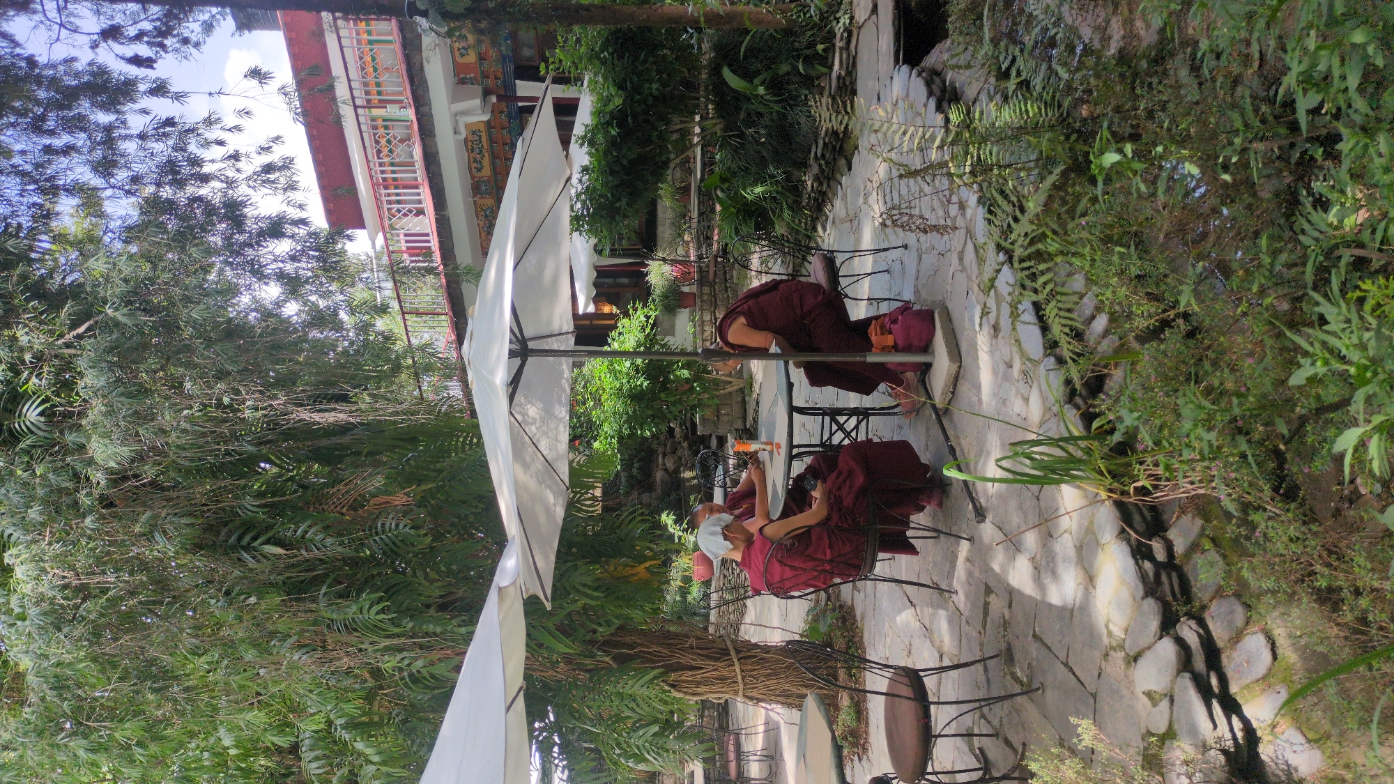 Two monks are seated at a table under a white umbrella in an outdoor patio. The monks are wearing traditional maroon robes. One is wearing a face mask. On the table between them is a beverage. The patio is paved with stone and bordered by lush greenery. The building in the background is multi-story and features red trim. The location may be in Fatehpur, India.