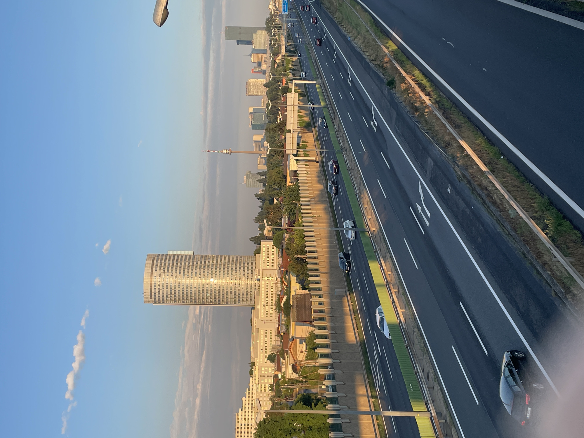 This is a wide shot photograph of a highway in Vienna, Austria, with a city skyline in the background. The sky is a clear blue with a few wispy white clouds. The sun is shining brightly, casting long shadows across the highway and illuminating the buildings in the distance. The highway has multiple lanes, with several cars driving on it.
In the foreground, the highway is bordered by a green sound barrier. Beyond the highway, there are several buildings of varying heights and architectural styles. The most prominent building is a tall, cylindrical skyscraper with a reflective facade. Other buildings include residential complexes and smaller commercial structures.
In the distance, the Vienna skyline is visible, featuring several skyscrapers, a tall radio tower, and other urban structures. The overall scene depicts a bustling urban environment with a major transportation artery cutting through it.