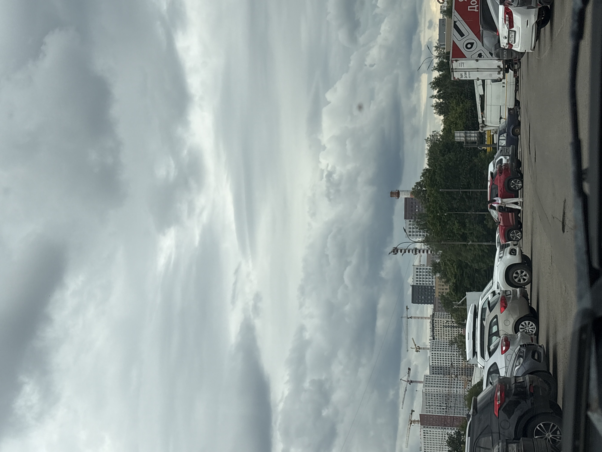 The image depicts a parking lot scene with several vehicles. In the foreground, a black SUV is parked on the left. To its right, a silver SUV and a white SUV are visible. In the middle ground, a red SUV is parked, and a woman in white clothing is standing near it. Further back, a white truck with red accents is parked. In the background, there are several tall buildings under construction with cranes. The sky is overcast with dense clouds. The location appears to be Tarasovka, Russia, as indicated by the context.