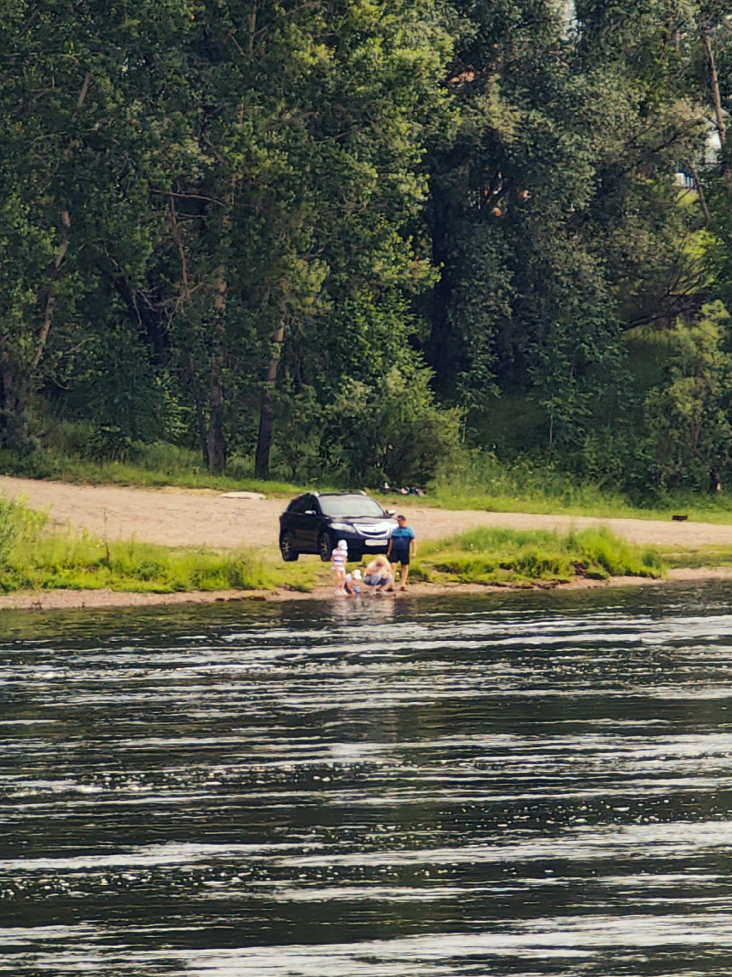 In Sosnovoborsk, Russia, a black SUV is parked on a sandy embankment next to a body of water. Several people are visible in the shallows. Two individuals, one appearing to be an adult male wearing a blue shirt and dark shorts, and another person (gender unclear) wearing a light-colored top and dark bottoms, are interacting near the water's edge. Closer to the SUV, two children, one in a striped outfit and another in a pink outfit, are also present in the water. The background is dominated by a dense forest of green trees. The water in the foreground exhibits ripples and reflections.