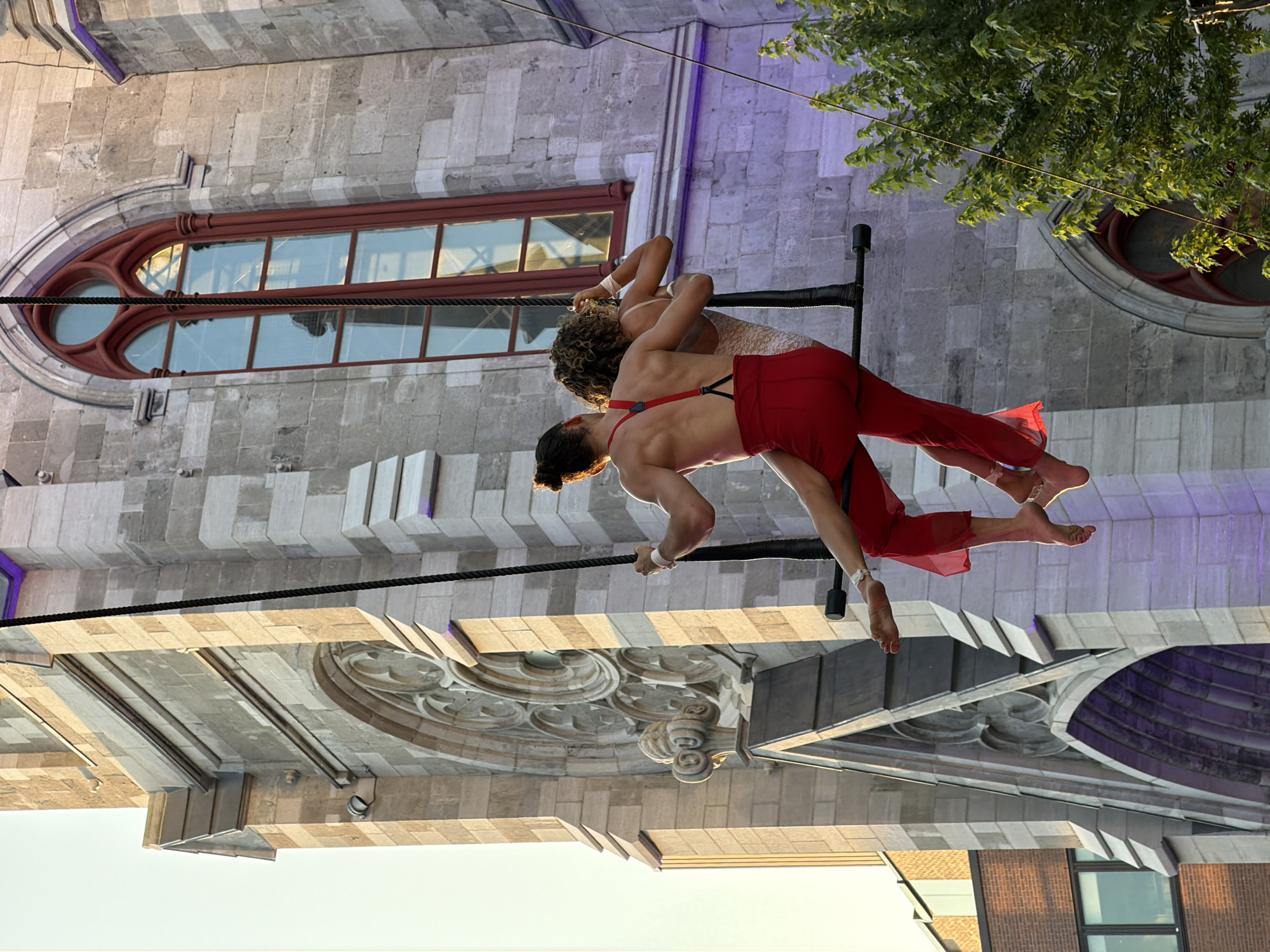 Two performers are suspended mid-air by a trapeze bar attached to ropes, against the backdrop of a large, stone Gothic-style building. The performer on the left, wearing red suspenders and red pants, is partially visible from the waist down, facing away from the camera. The performer on the right, wearing a light-colored, textured outfit, is embracing the performer on the left. The building features a tall, arched window with red framing and stained glass reflections, and intricate stone carvings. The scene appears to be in Montreal, Canada, likely near a historic church or cathedral. A tree with vibrant green foliage is visible on the right side of the image.