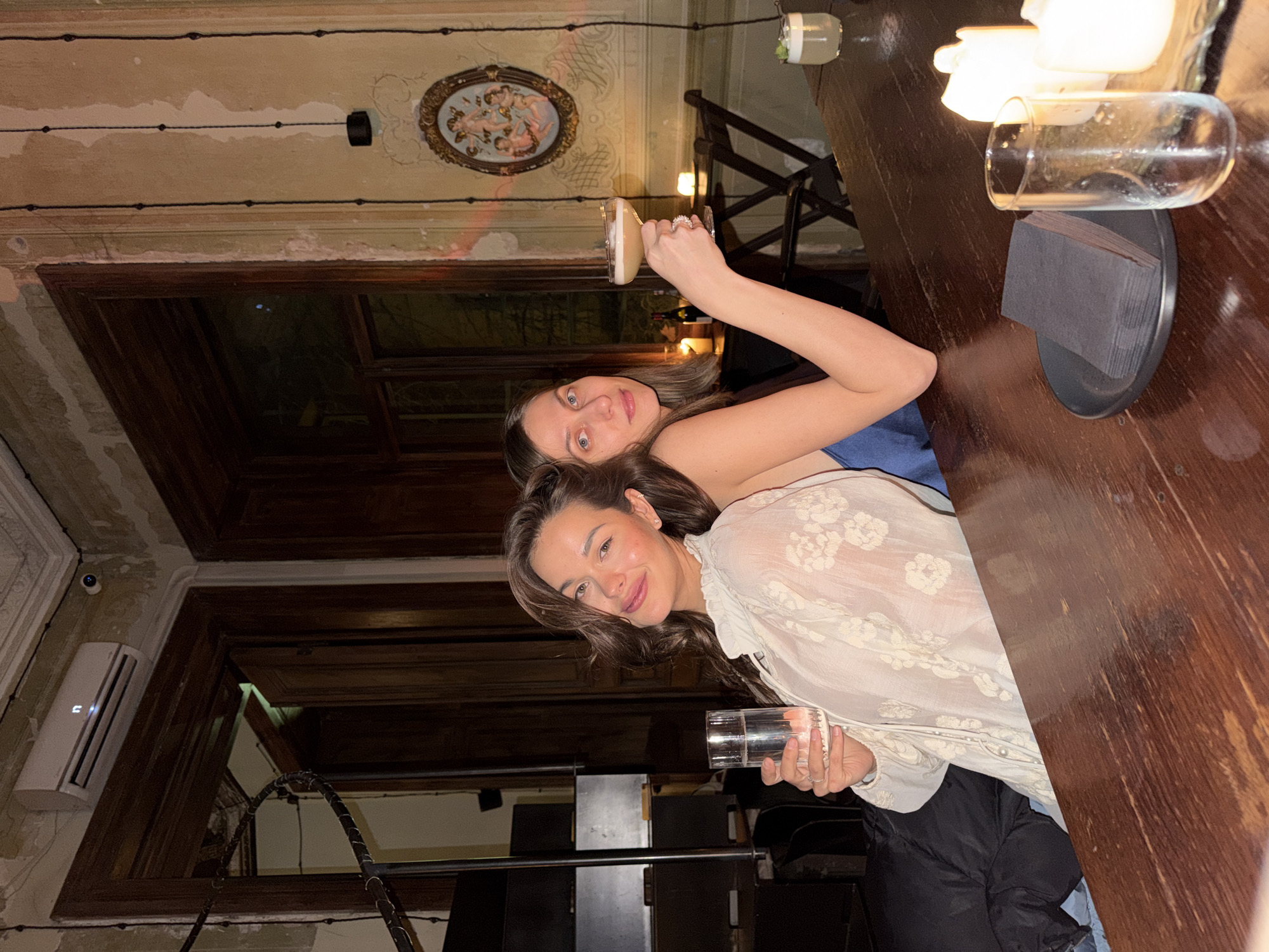 Two women are seated back-to-back at a dark wooden bar or table within a dimly lit establishment in Tbilisi, Georgia. The woman on the left, with dark wavy hair, wears a light-colored, sheer blouse with floral embroidery and holds a clear glass of water in her right hand, smiling directly at the camera. The woman on the right, with straight brown hair, wears a dark top with a blue strap and holds a frothy cocktail in a coupe glass in her left hand, gazing towards the camera. On the table, to the right, are three white pillar candles (two lit), an empty clear glass tumbler, and a stack of black napkins on a small black plate. A short glass with a light-colored drink and green garnish is visible further down the table. The background features aged, light-colored walls with peeling paint, dark wooden doorframes, and an ornate ceiling with molding. Above a doorframe to the left, a white air conditioning unit with an illuminated 'n' is mounted, accompanied by a small black security camera. To the right, an oval-framed painting depicting cherubs hangs on the wall, near a window with dark frames and a small table with additional lit candles and a bottle. A dark spiral staircase railing is partially visible on the left.