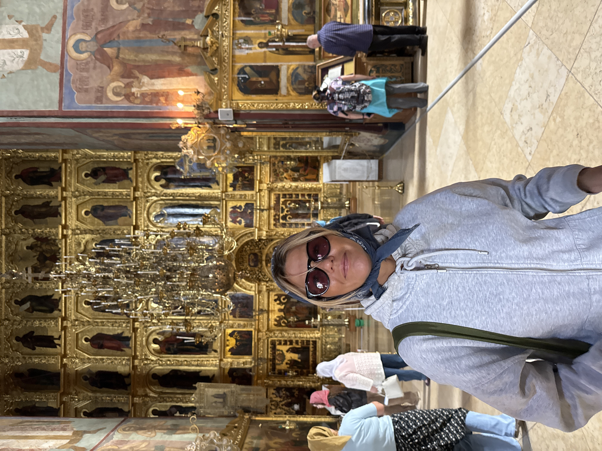 This image captures a candid moment inside the Trinity Lavra of Saint Sergius, a historic monastery located in the city of Sergiev Posad, Russia. In the foreground, a woman with blonde hair and sunglasses is looking at the camera. She is wearing a grey hooded sweatshirt and a blue headscarf. Behind her, other visitors are seen browsing the ornate interior of the church. The background is dominated by a large iconostasis, a wall of religious icons, and a gilded chandelier. The floor is made of light-colored tiles. The scene is illuminated by natural light and the warm glow of candles.