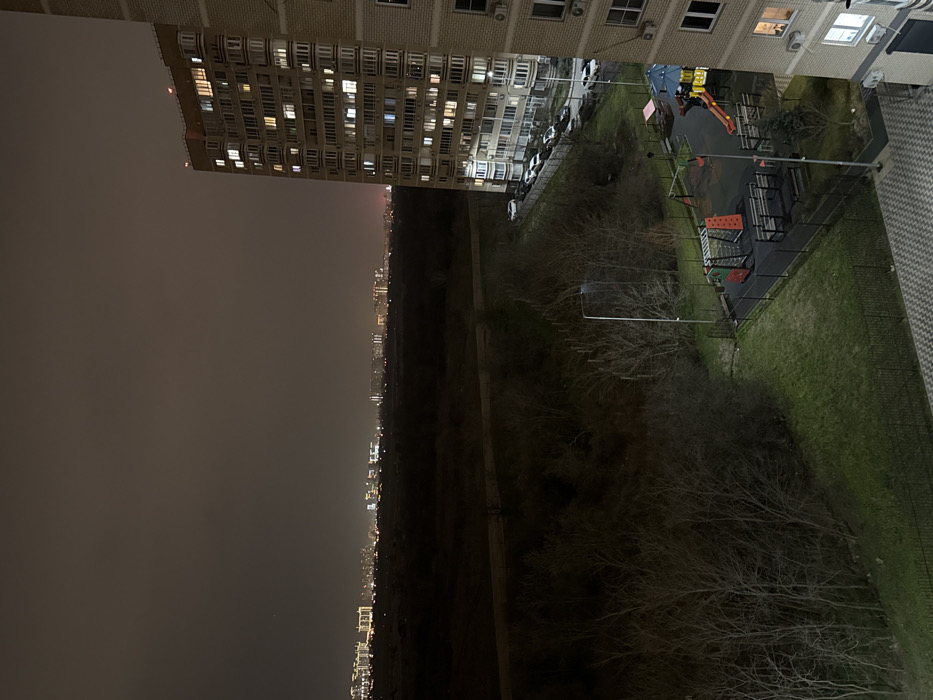 This night-time image captures an urban landscape adjacent to undeveloped land in Krasnodar, Russia, viewed from an elevated position. The dark, overcast sky features a faint reddish glow along the distant horizon, indicative of light pollution from the sprawling city lights that define the far background.
A significant portion of the mid-ground consists of dark, undeveloped terrain covered by numerous bare, deciduous trees and shrubs. A faint, unpaved track traverses horizontally through this natural area.
To the right, a multi-story residential building with a light-colored facade stands prominent. Many of its windows are illuminated, and balconies are visible on multiple floors. Air conditioning units are mounted on its exterior. Adjacent to the building, a paved area serves as a parking space for approximately eight white passenger vehicles.
In the central foreground, a fenced children's playground features various colorful equipment, including red and blue slides, climbing structures, and multiple benches. The playground ground surface appears dark and possibly rubberized. Several tall streetlights illuminate this area, casting distinct shadows. Green grass and more bare trees occupy the immediate left foreground, transitioning into the larger natural area. The bottom edge of the image reveals a patterned tiled surface, indicating the observation point is from a balcony or similar structure. No individuals or animals are visible within the frame.
