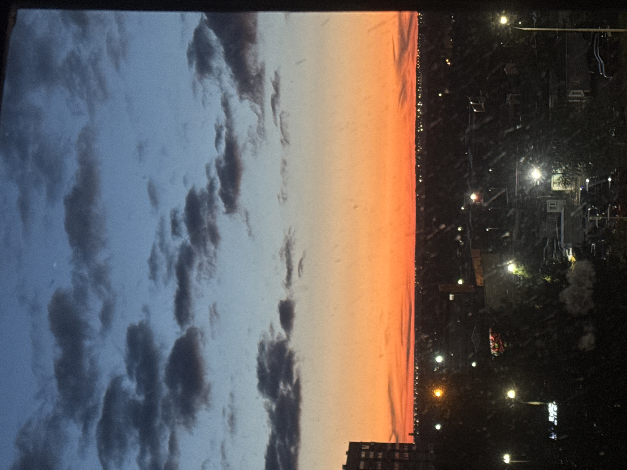 This image captures a high-angle view of a city or urban landscape at dusk or sunset, likely taken from a window. The scene is horizontally oriented but the provided image is rotated 90 degrees clockwise.
In the upper portion, the sky transitions from a soft blue with scattered, textured dark grey clouds to a vibrant orange and pink hue closer to the horizon, indicating the setting sun. The horizon line runs across the middle of the scene, separating the colorful sky from the darker cityscape below.
The lower portion of the image reveals the city at night, plunged into darkness but illuminated by numerous artificial lights. These lights appear as small, bright dots scattered throughout the dark expanse, suggesting streetlights, building lights, or distant vehicles. Silhouettes of buildings, including one distinct multi-story structure, are visible against the brighter horizon, while other faint structural outlines and dark masses that could be trees or other foliage emerge from the darkness among the lights.
There is no specific activity taking place, rather it is a serene landscape view capturing the transition from day to night. Notable details include the vivid sunset colors in the sky and the glowing artificial lights of the city below. The image appears to have been taken through a surface, possibly a window, as there are visible specks, dust, or smudges, particularly noticeable in the darker areas and on the glowing lights. No people are visible, and there is no discernible text in the image. Given the provided context, the location is Izhevsk, Russia.