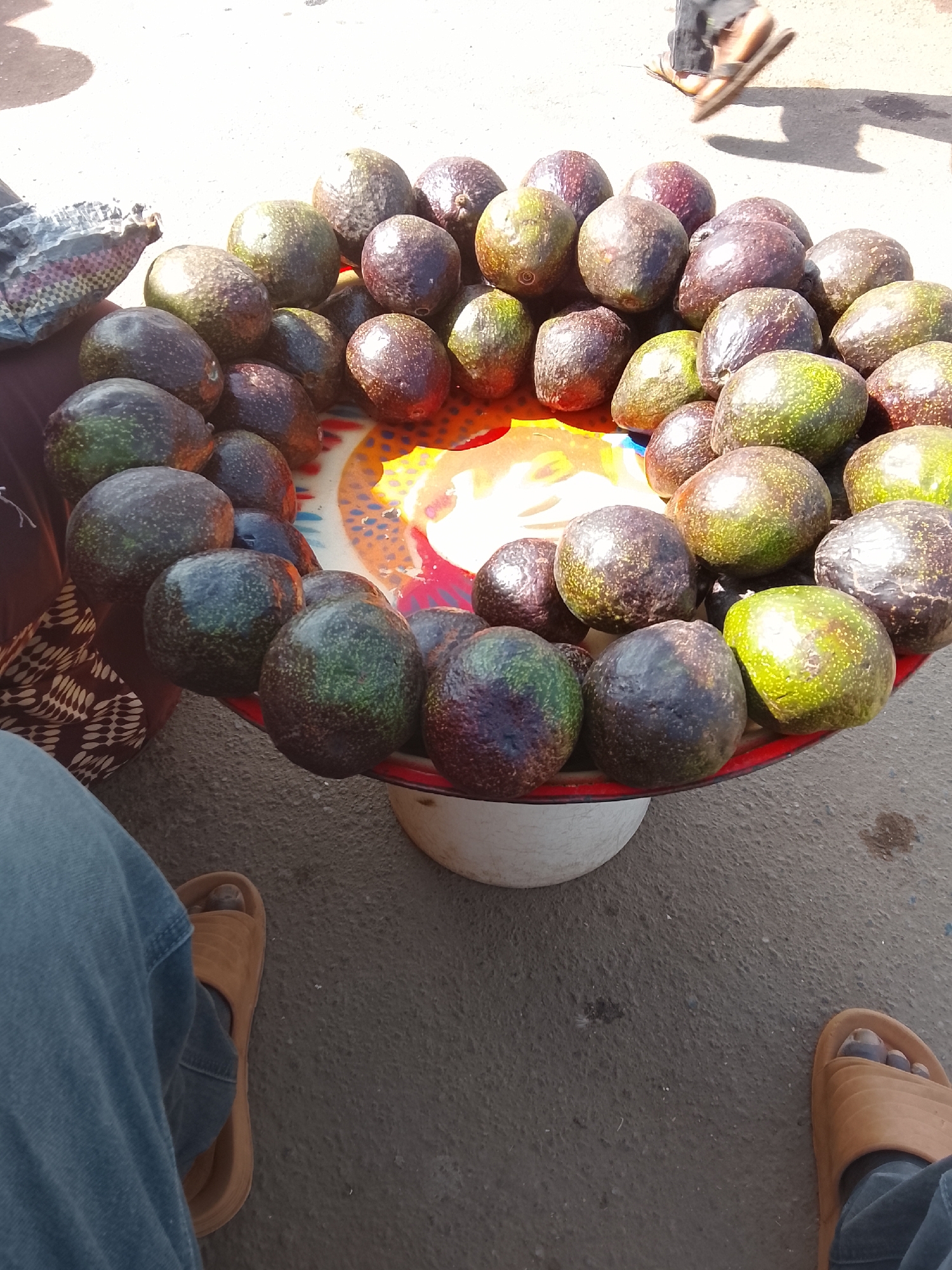 A street scene in Gwong, Nigeria shows a vendor selling avocados. The avocados are arranged in a single layer on a colorful patterned tray, which is balanced on a white bucket or container. The avocados are various shades of green and dark purple. Parts of two people are visible: One person is seated near the tray wearing blue jeans and tan sandals, and another person is walking in the background, also wearing tan sandals. The ground is paved.