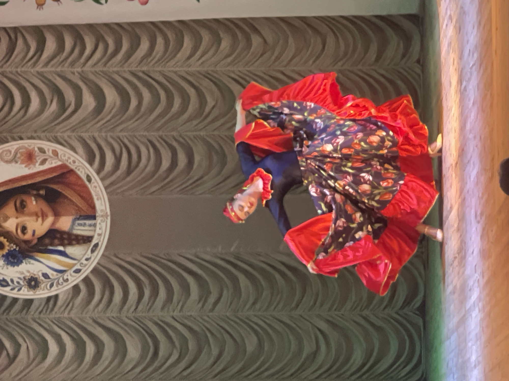 A young woman in traditional dress dances on a wooden stage in front of a backdrop of draped fabric. She wears a dark blue bodice with red trim and a wide, multi-layered skirt featuring a floral pattern of red and pink flowers on a dark background, with a bright red underskirt. Her hair is pulled back and adorned with a red headband. A circular painting of a young woman with braided hair and floral elements is visible above the draped backdrop. The stage is lit from above, casting shadows on the wooden floor. The context suggests a performance or cultural event in the city of Horozhuli, Ukraine.