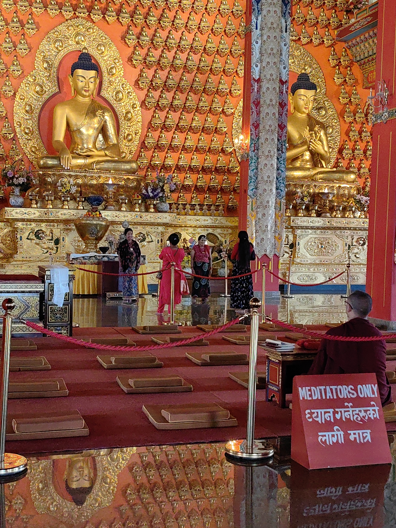 This image shows the interior of a Buddhist temple in Parsa, Nepal. The temple features two large golden Buddha statues seated against a wall covered with numerous smaller golden Buddha figures. In the foreground, there are rows of meditation cushions laid out on a red carpet. Several people are present in the scene. Two women in traditional attire stand near the statues, while a Buddhist monk in maroon robes is seated in the lower right corner, facing away from the camera. A red sign with white lettering, reading "MEDITATORS ONLY" in English and Nepali script, is placed on the floor. The floor is highly reflective, showing a mirror image of the temple's decor and a reflection of a Buddha's face.