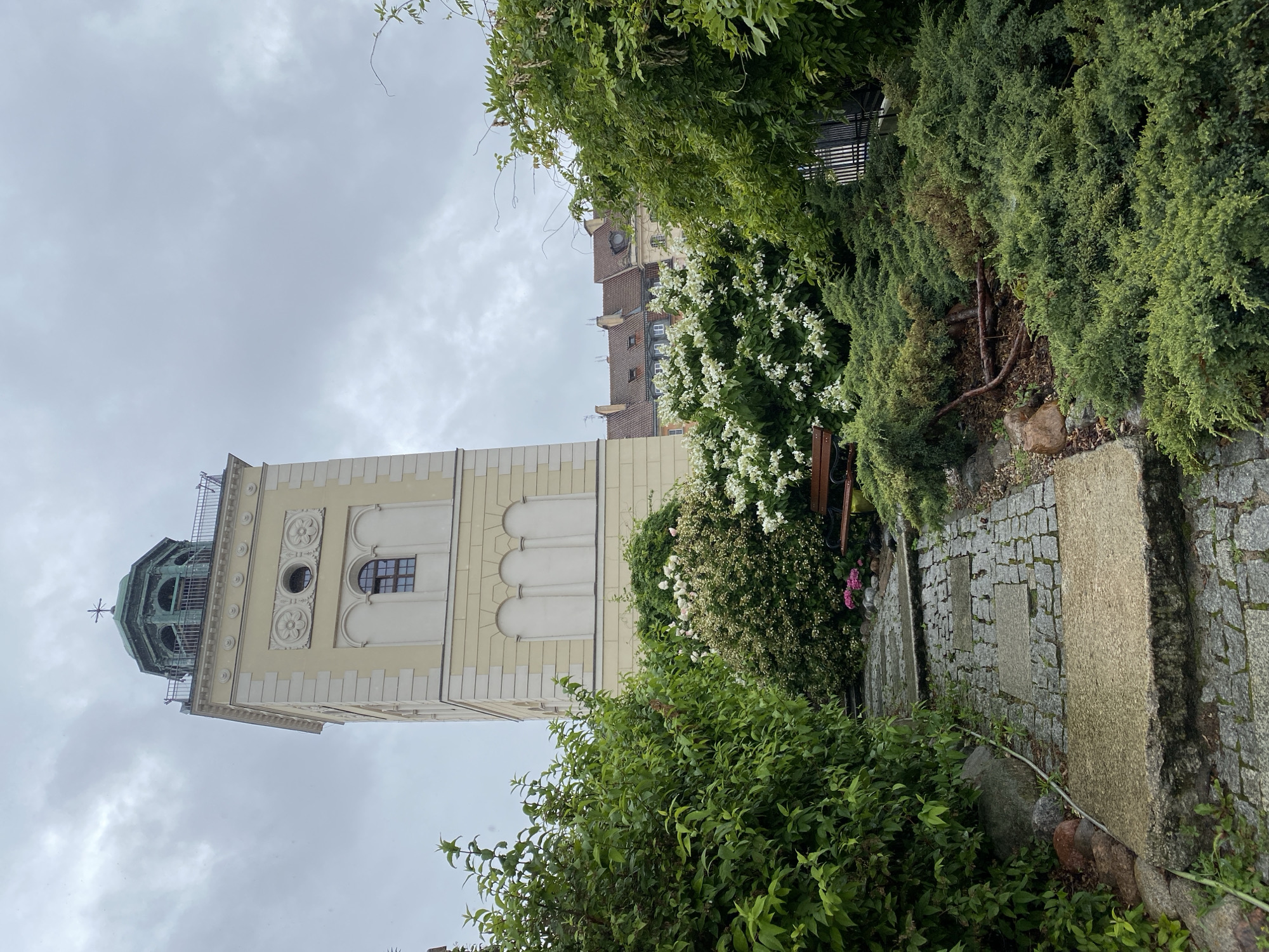 The image shows a view of a historical tower in Warsaw, Poland, partially obscured by lush green foliage. The tower, painted a pale yellow, has a distinctive green, domed cupola topped with a cross-like ornament. Several arched windows and decorative circular elements adorn its façade. Adjacent to the tower, a building with a red-tiled roof and dormer windows is visible. The foreground is dominated by dense greenery, including flowering bushes with white blossoms and tall evergreen shrubs, creating a sense of a garden setting. A stone pathway, laid with cobblestones and interspersed with larger rectangular stones, leads through the greenery. The sky is overcast, suggesting a cloudy day, and the overall lighting is soft. There is no visible text in the image. The scene evokes a peaceful, somewhat enclosed atmosphere, with the natural elements softening the architectural lines of the tower and buildings.