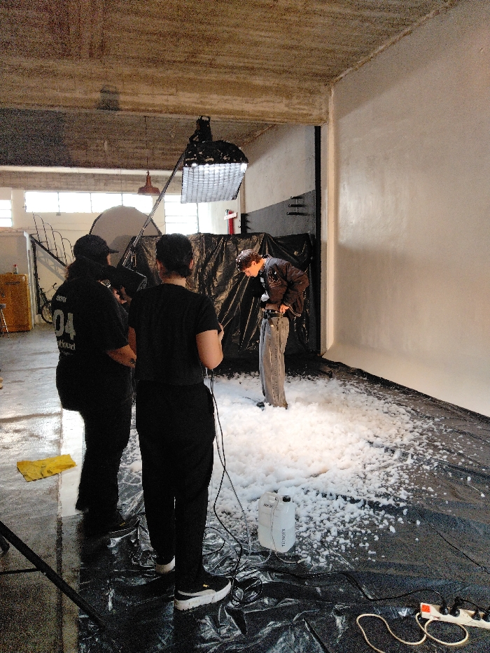 A behind-the-scenes shot shows a film or photo shoot in progress. Two crew members in black stand watching a model adjusting their belt in a pile of white foam. A boom light and reflector are visible above the set, which is covered in a black plastic sheet. A white container labelled "TC.MFX" with attached wiring sits near the crew members, and an extension cord is plugged into the ground. Windows are seen in the background, providing natural light in addition to the artificial lighting setup.