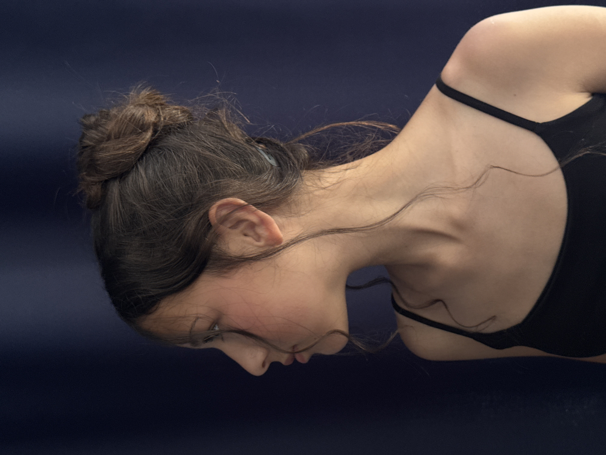 The image displays a profile portrait of a young person with dark hair styled in a messy bun. The person is looking to the left, with a slightly parted mouth. They are wearing a black tank top. The background is a solid dark blue fabric, which appears to be a curtain or backdrop. The lighting highlights the contours of the person's face and neck. There are no discernible elements in the image that indicate a specific location in Sochi, Russia, or any urban setting.