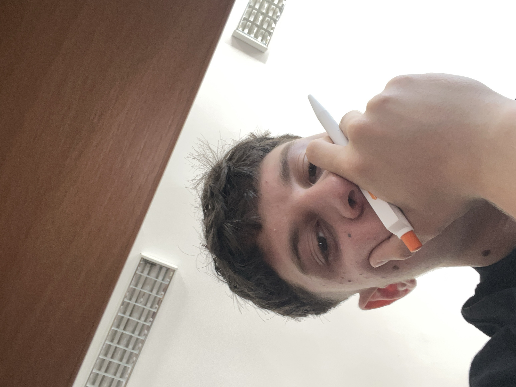 A low-angle, close-up selfie captures a young male with dark, short, tousled hair. He is looking down at the camera, his face partially obscured by his hand holding a white and orange object, possibly a pen or stylus, to his cheek. His skin appears to have some blemishes. He is wearing a dark t-shirt. In the background, there is a white ceiling with fluorescent light fixtures and a wooden soffit. The scene is indoors and does not provide any specific visual cues to indicate a location within the city of Zlatograd, Bulgaria.