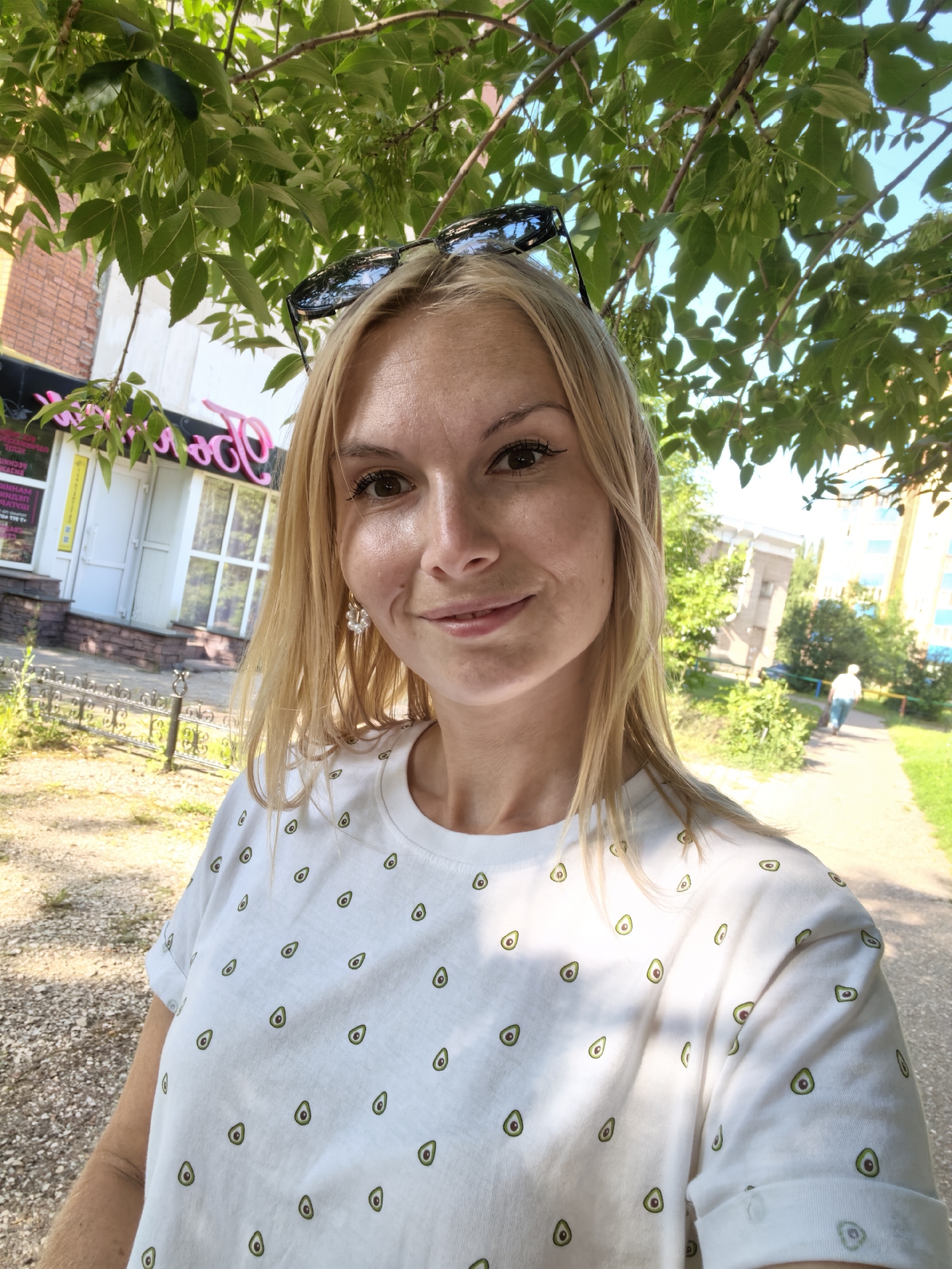 A young woman with blonde hair and sunglasses on her head is taking a selfie outdoors. She is wearing a white t-shirt with a pattern of avocados. In the background, there is a building with pink lettering, trees, a sidewalk, and a person walking away. The scene appears to be in a city, likely Sterlitamak, Russia, given the context. The lighting suggests it is daytime and sunny.