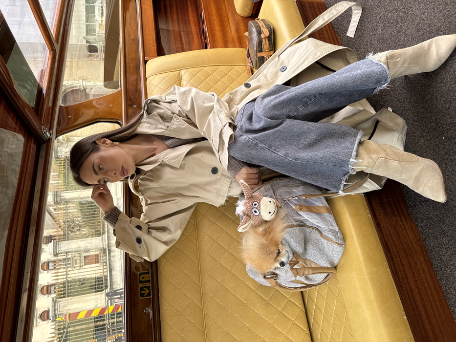 A woman is seated inside a water taxi on a canal in Venice, Italy. She wears a light beige trench coat, a grey sweater, wide-leg blue jeans with frayed hems, and light beige pointed-toe boots. Her right hand rests on her forehead as she gazes towards the left side of the frame. Beside her on the quilted yellow upholstered seat, a grey duffel bag holds a small, fluffy orange-brown dog, with a crocheted donkey plush toy attached to the bag's handle. Further to her right on the seat, a small, brown Louis Vuitton hard-shell suitcase is present. The boat's interior features varnished wooden panels and structural elements. Through the window, classical Venetian architecture is visible, including a stone balustrade with pillars, buildings, and a distinctive red and yellow striped pole on the water's edge.