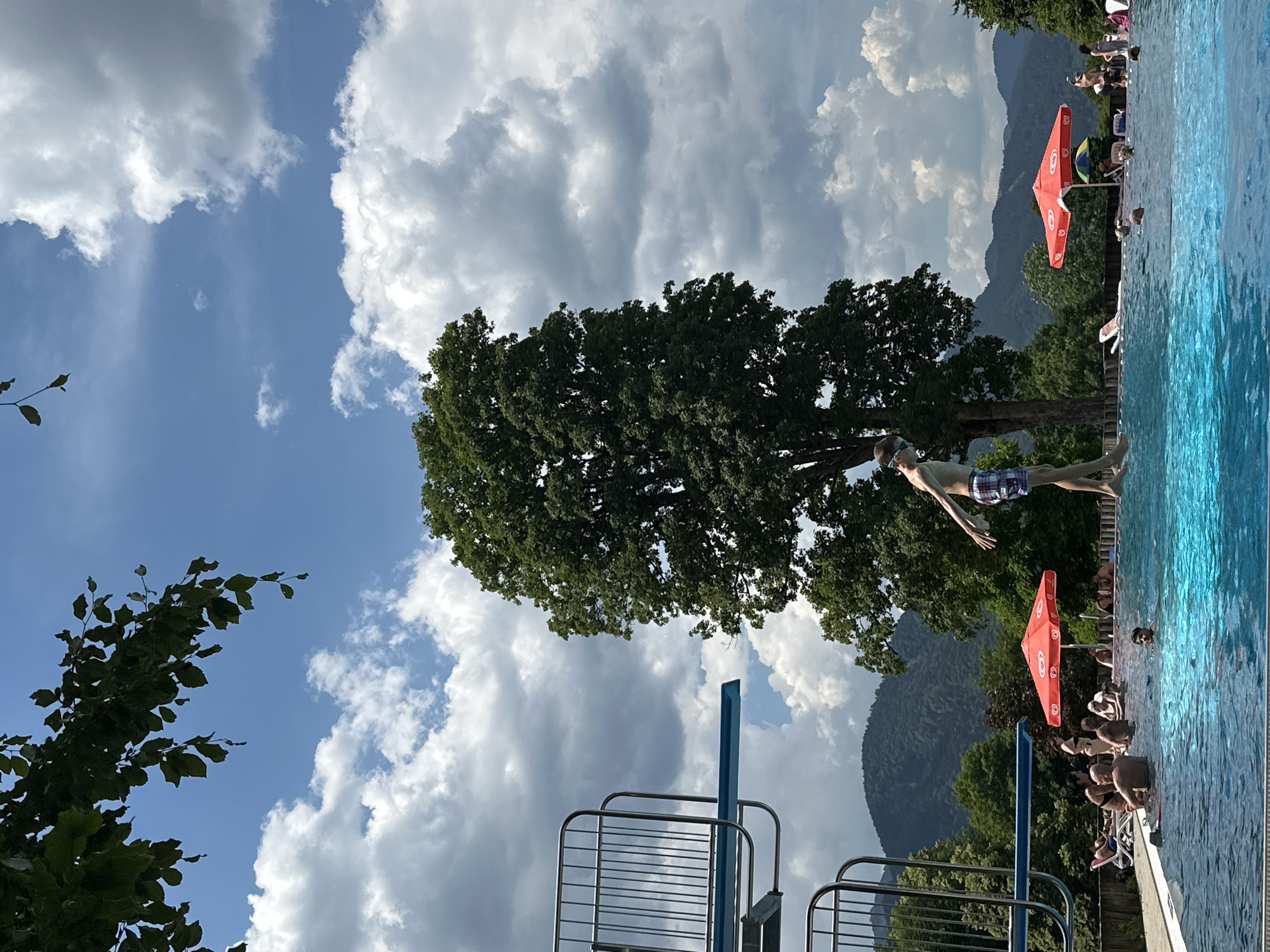 The image depicts a public swimming pool with a diving board and several people enjoying a summer day. In the foreground, a young person in plaid shorts is captured mid-air, having jumped from the diving board. The pool water is a vibrant blue. Surrounding the pool, people are swimming, sunbathing, and relaxing under red umbrellas. Lush green trees and mountains form the backdrop under a partly cloudy sky. The scene appears to be located in Oberammergau, Germany, as suggested by the presence of a diving tower and the scenic mountain landscape typical of the region.