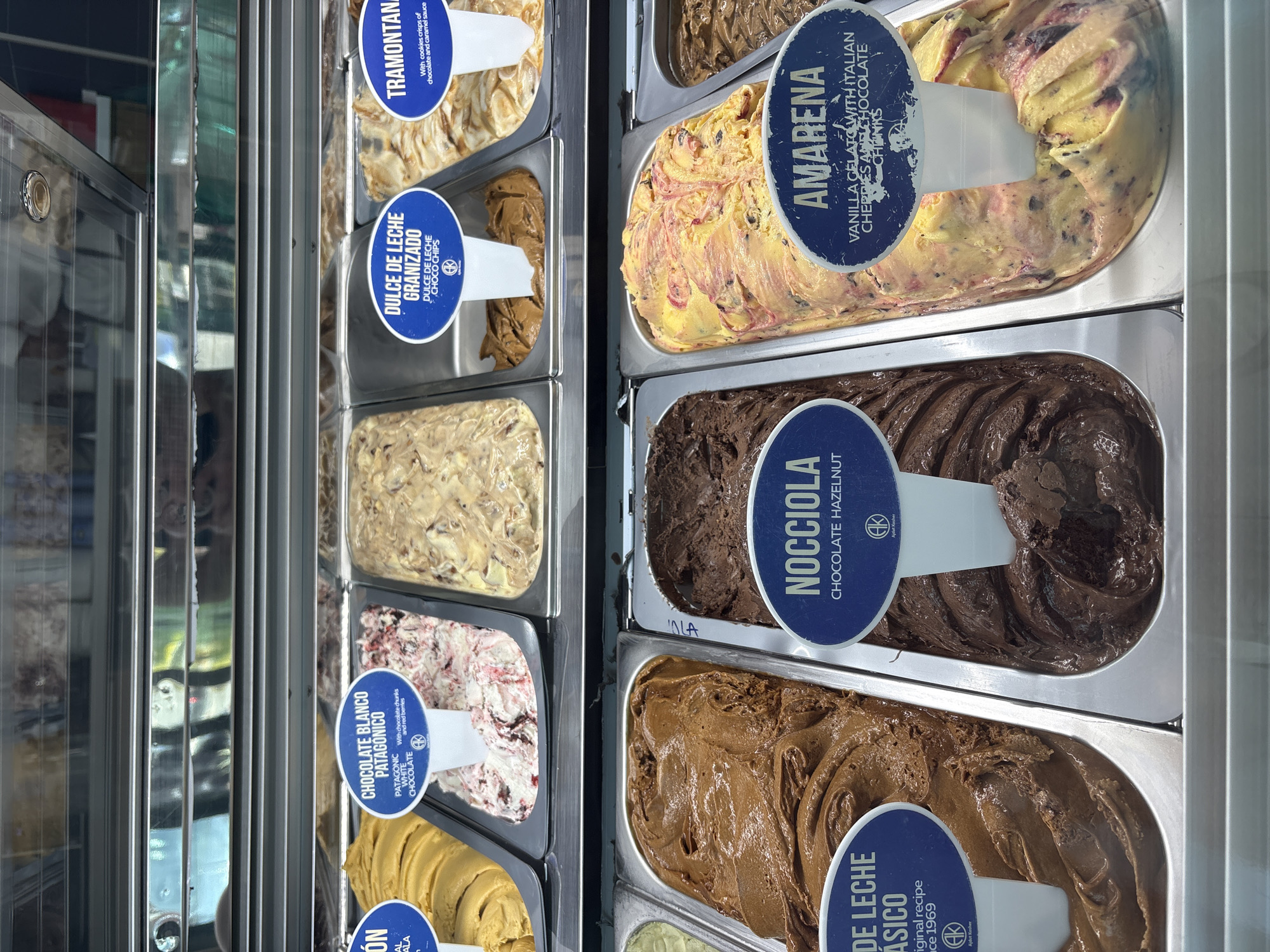The image displays a detailed, downward-angled view of a commercial gelato display case. The case contains eight visible stainless steel trays, each holding a different flavor of gelato. Above each flavor, a blue oval-shaped sign with white text identifies the product. A chrome-finished strip runs horizontally above the gelato, reflecting ambient lighting and indistinct shapes from the environment.
In the top row, from left to right, a portion of a yellow-orange gelato is visible. This is followed by a white gelato labeled "CHOCOLATE BLANCO PATAGONICO" (Patagonic White Chocolate) containing red swirls and dark specks, described as having chocolate chunks and red berries. Adjacent to this is an unlabeled light brown, chunky gelato. Next, a light brown gelato labeled "DULCE DE LECHE GRANIZADO" (Dulce de Leche Choco Chips). The final visible flavor in this row is "TRAMONTANA," described as having cookie crisps of chocolate and caramel sauce.
In the bottom row, from left to right, a rich brown gelato labeled "DULCE DE LECHE CLASICO" (Dulce de Leche Classic), noted as an original recipe from 1969. Following this is a dark brown gelato labeled "NOCCIOLA" (Chocolate Hazelnut). Next is a yellow gelato with prominent red swirls and dark chunks, labeled "AMARENA" (Vanilla Gelato with Italian Cherries and Chocolate Chunks). A dark brown gelato is partially visible at the far right.
Several labels prominently feature an "AK" logo, indicative of potential Kosher certification. No individuals are present in the frame. The specific location of this establishment within Huixquilucan de Degollado, Mexico, cannot be determined from the visual information provided.